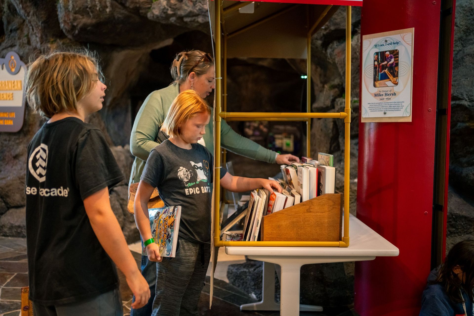 Three people stand at a red kiosk, browsing books on a table.