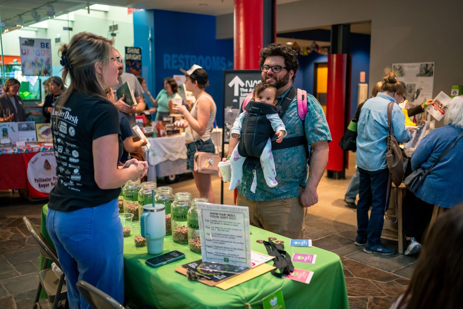 People chatting at a colorful indoor market booth with a green table and displayed products.