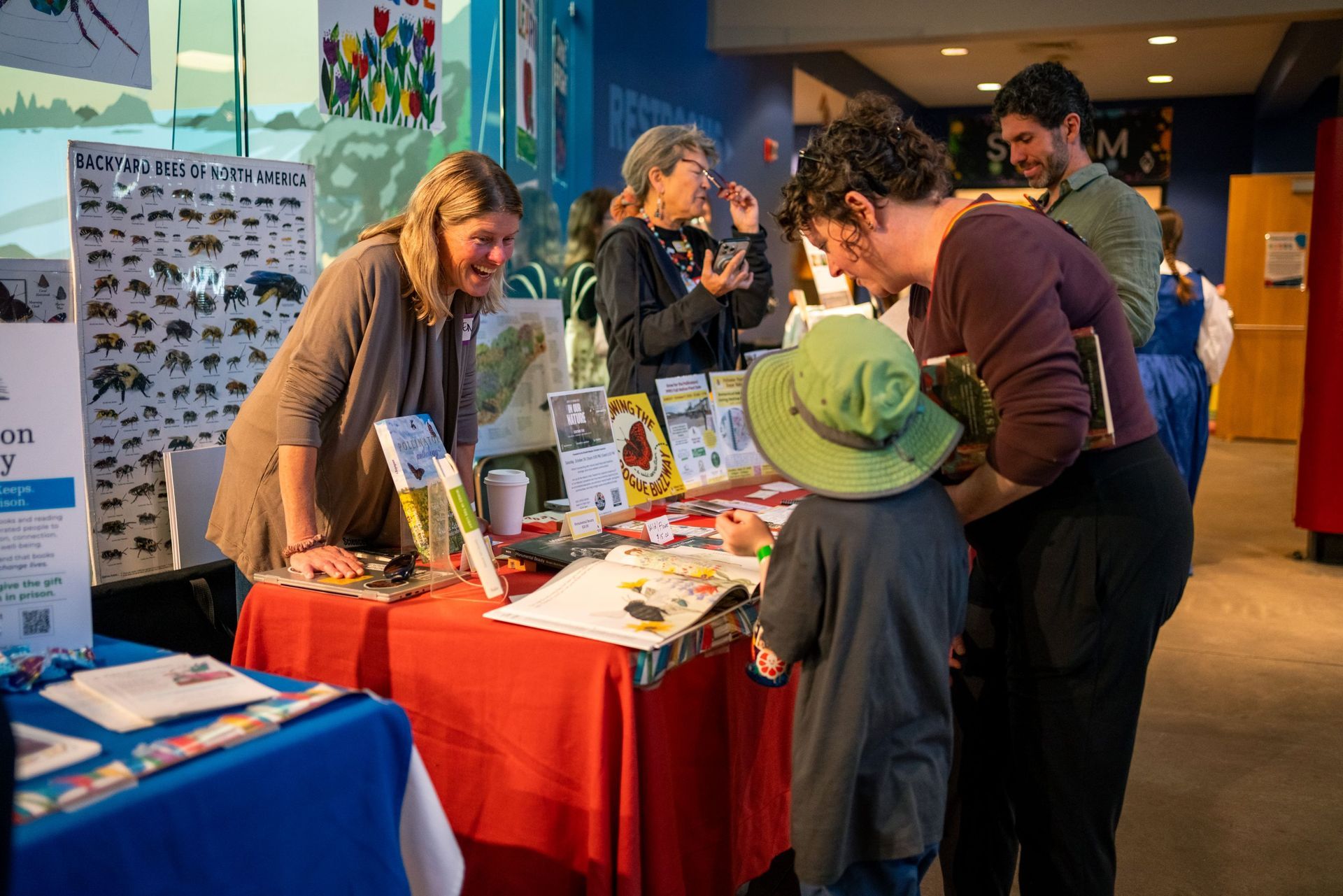 People at a table display in a hallway, with brochures and a child in a green hat looking at items.