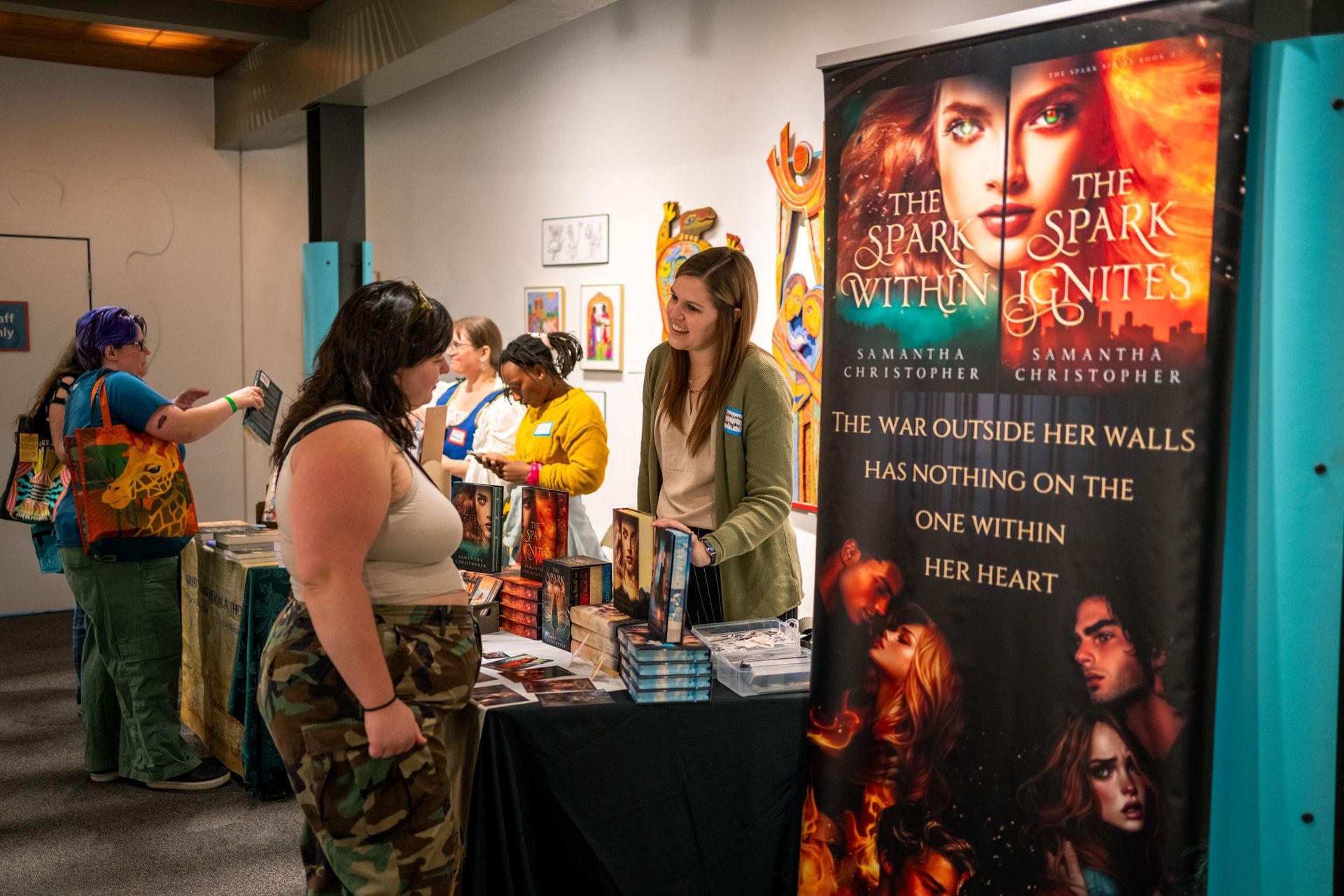 Book fair booth with a large fantasy novel poster and people browsing signed books.