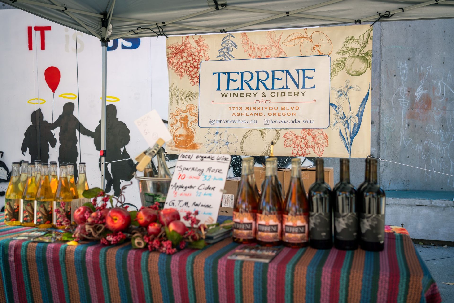 Market stall with bottles, produce, and a Terrene sign on a striped tablecloth.