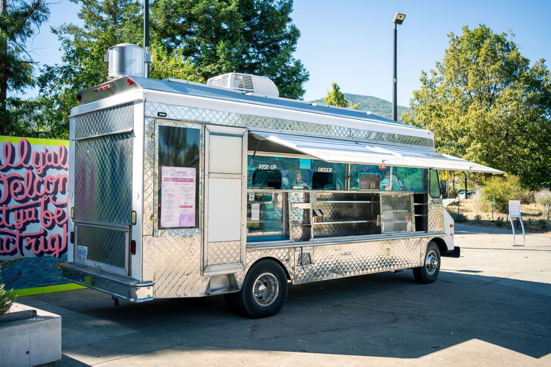 Silver food truck parked outdoors under blue sky, with a service window and striped awning.