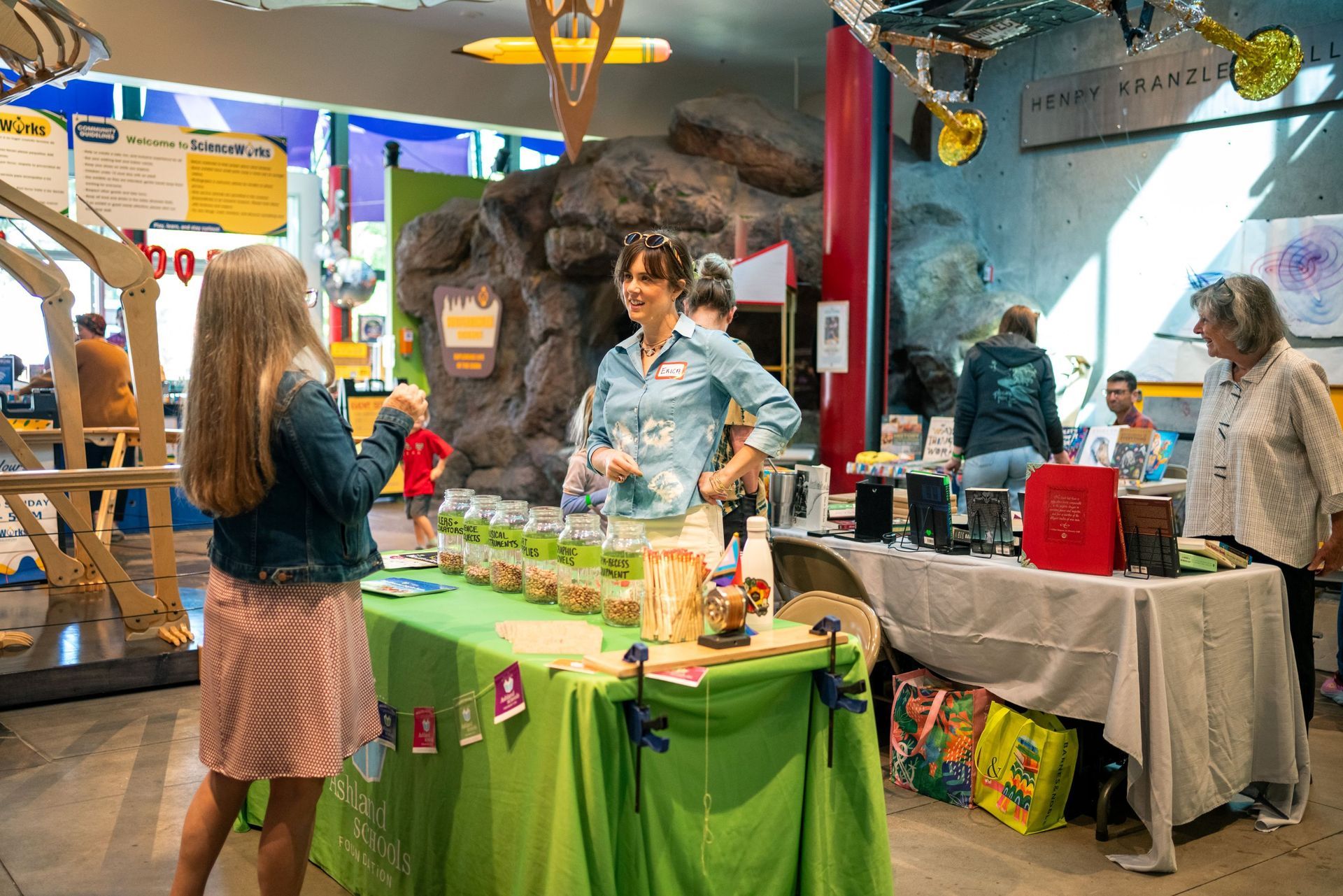 People at a green-clothed booth with samples and displays in an indoor market or fair.
