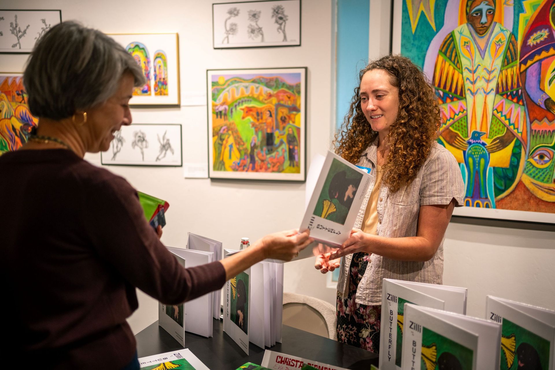 booth with colorful framed paintings in the background; a woman hands a book to a smiling guest.