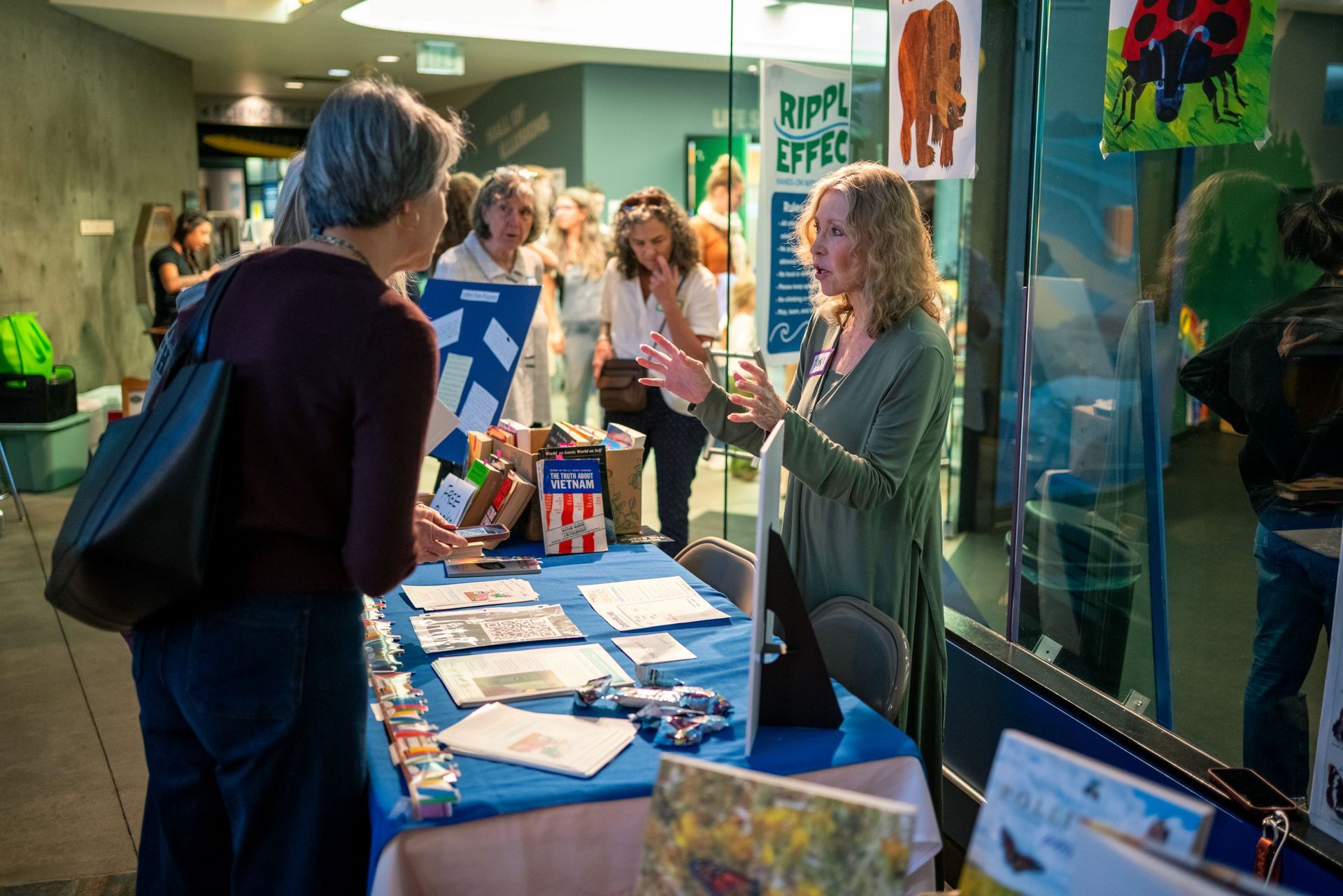 People talking at a blue booth indoors, with brochures and colorful posters displayed.