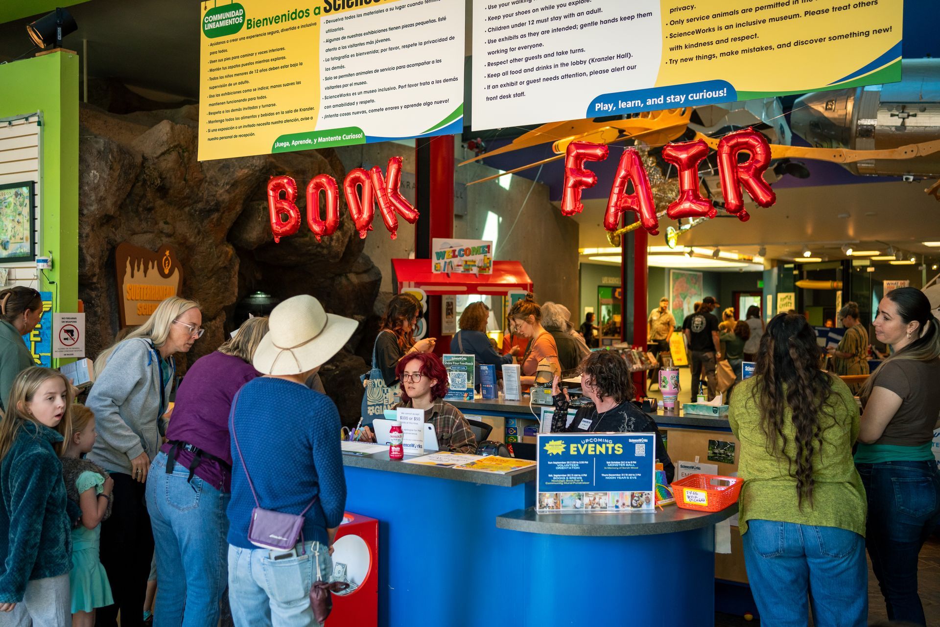 Book fair with people browsing at a blue counter under a red “BOOK FAIR” sign.