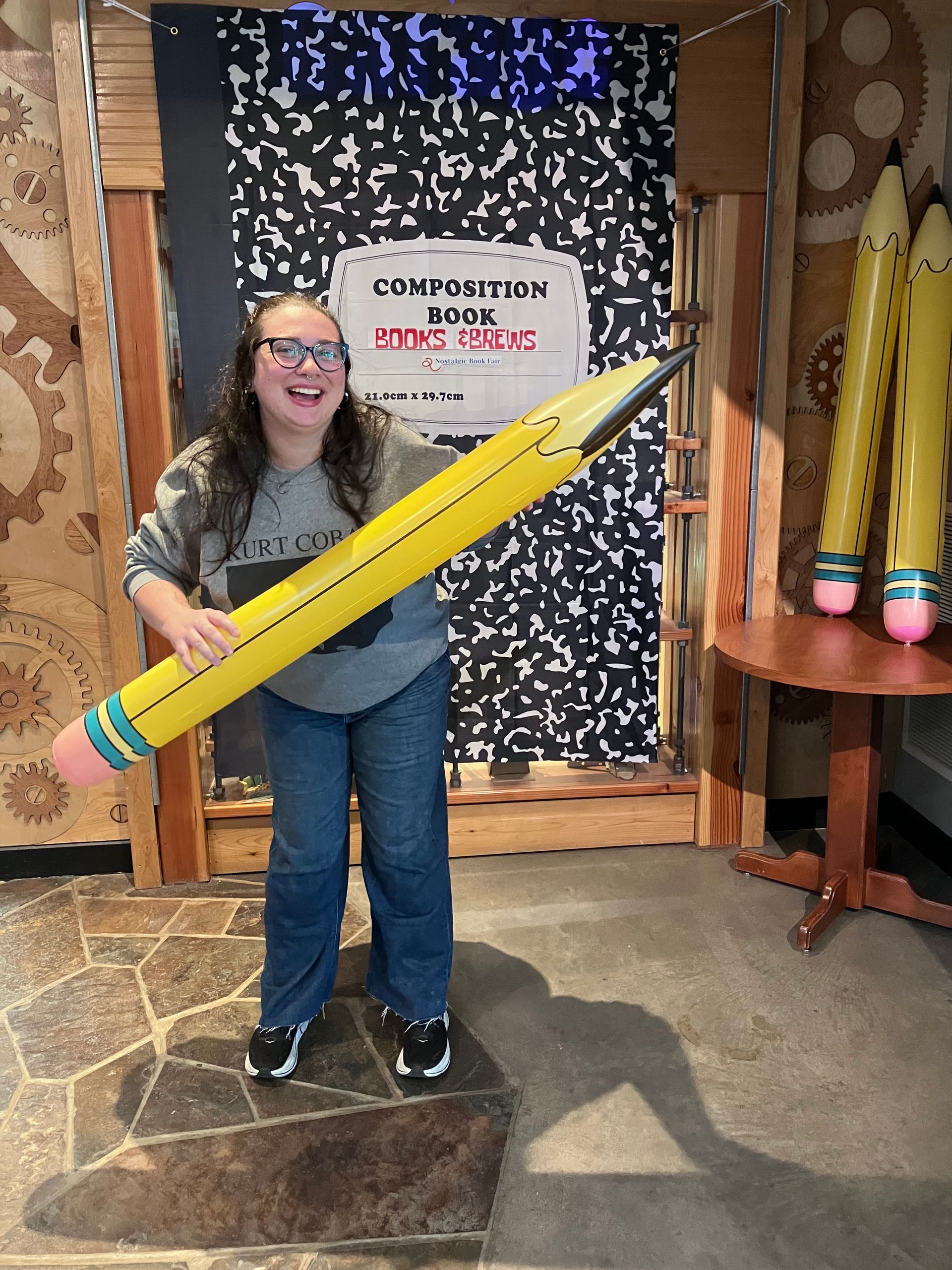 Person holding a giant yellow pencil in a museum display area with a sign behind them