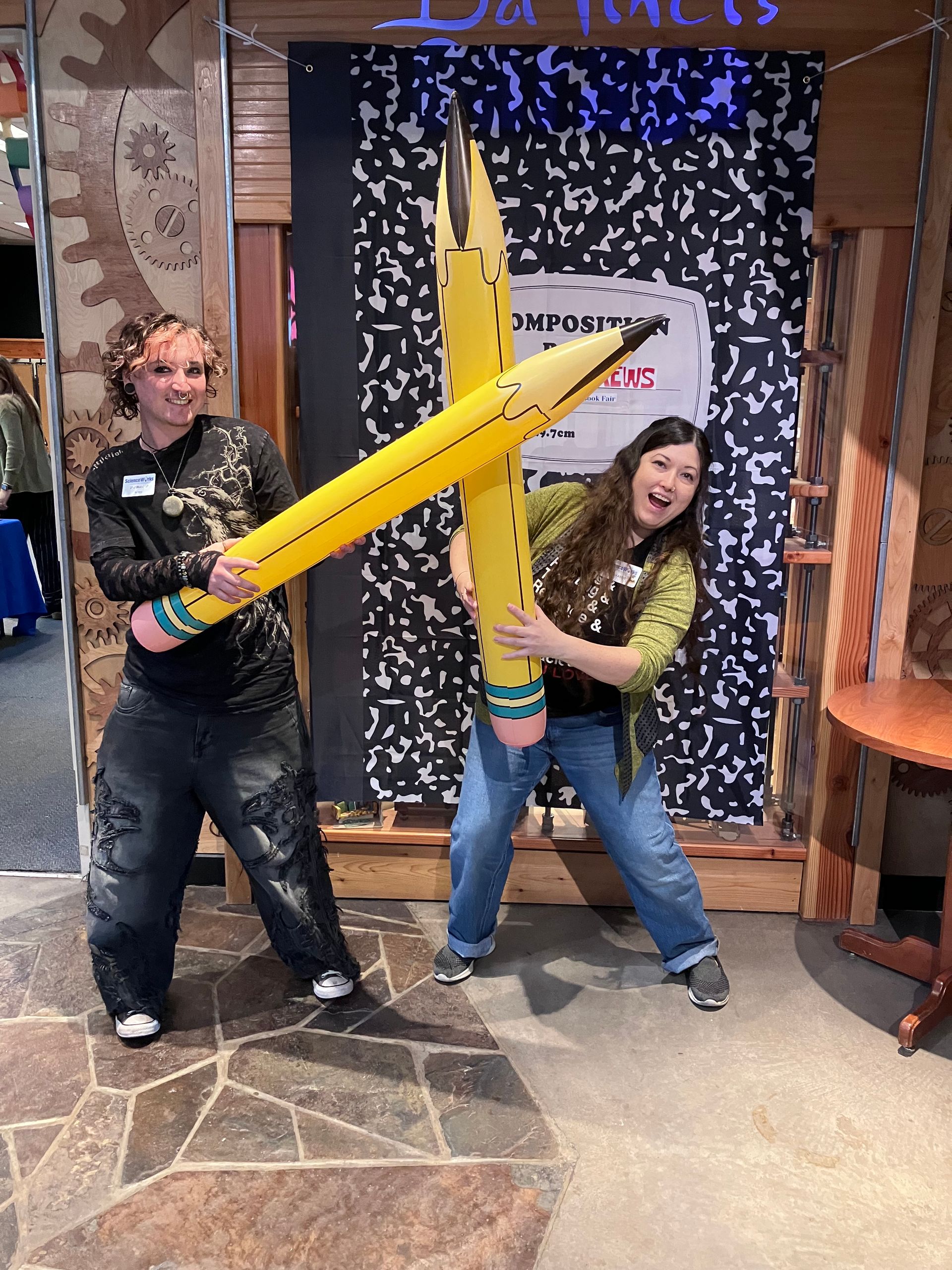 Two people posing with oversized yellow inflatable pencils in a playful setting.
