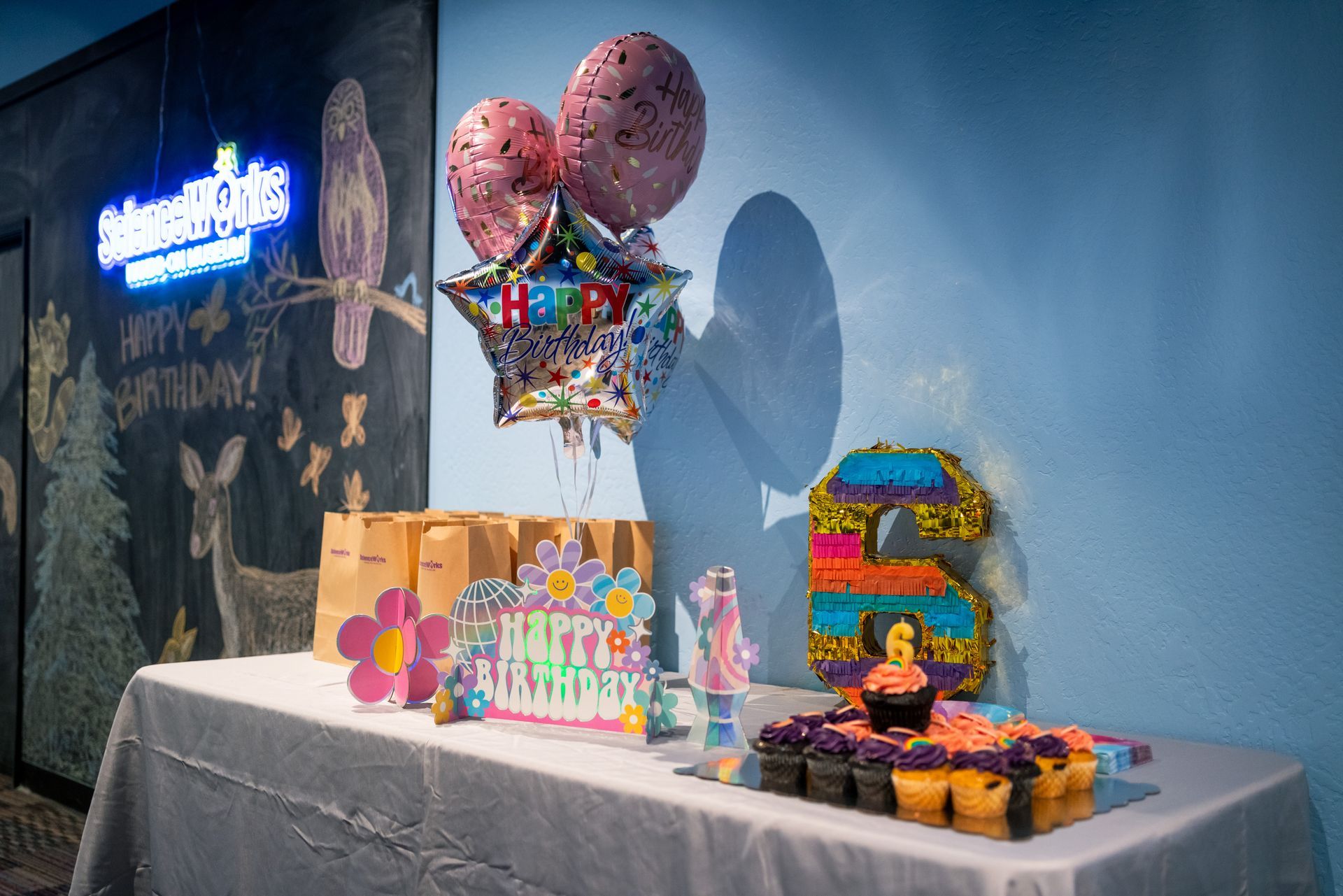 Birthday party dessert table with balloons, cake, gifts, and colorful treats against a blue wall
