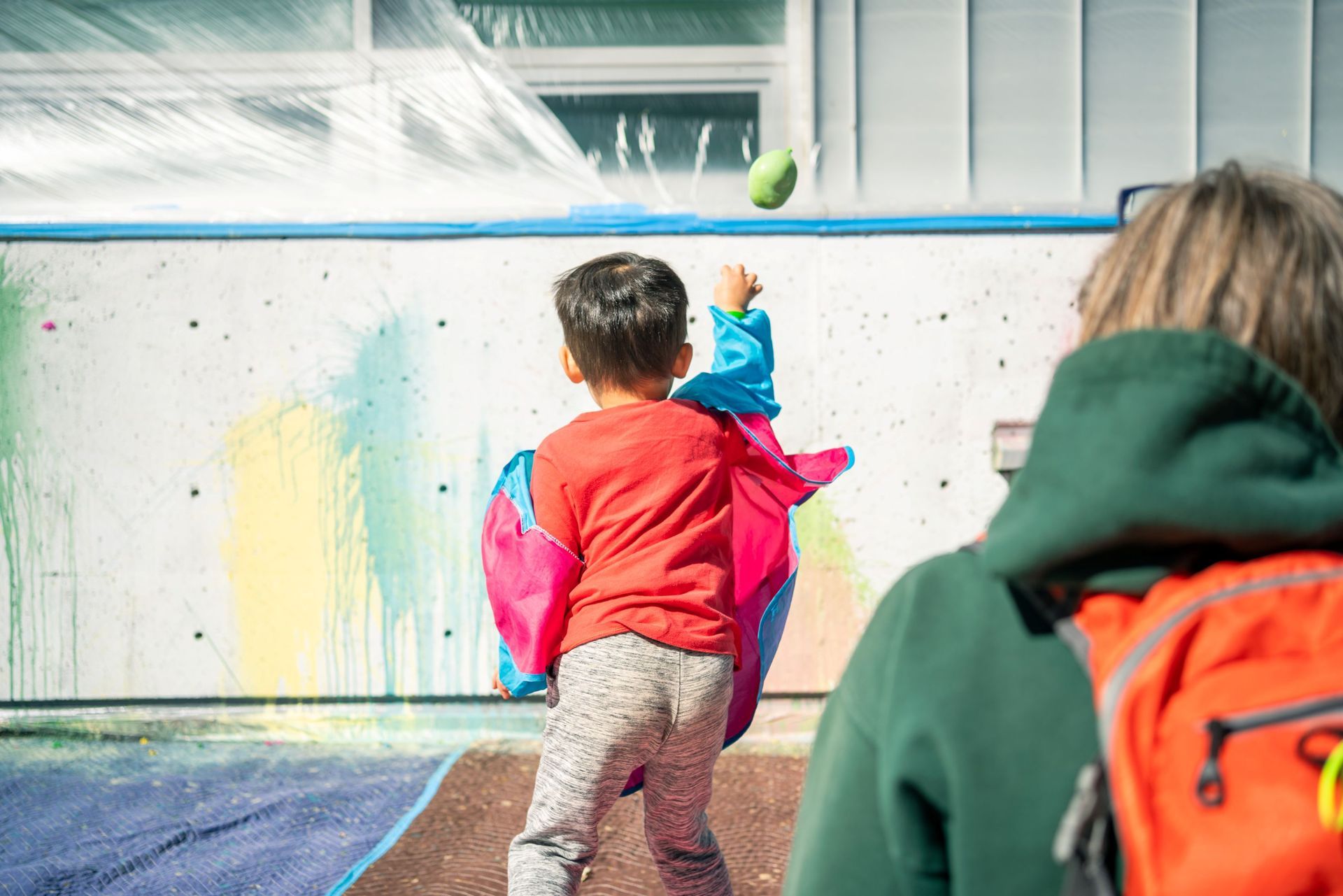 Child in red shirt throws a paint balloon at a paint-splattered wall while another person watches nearby