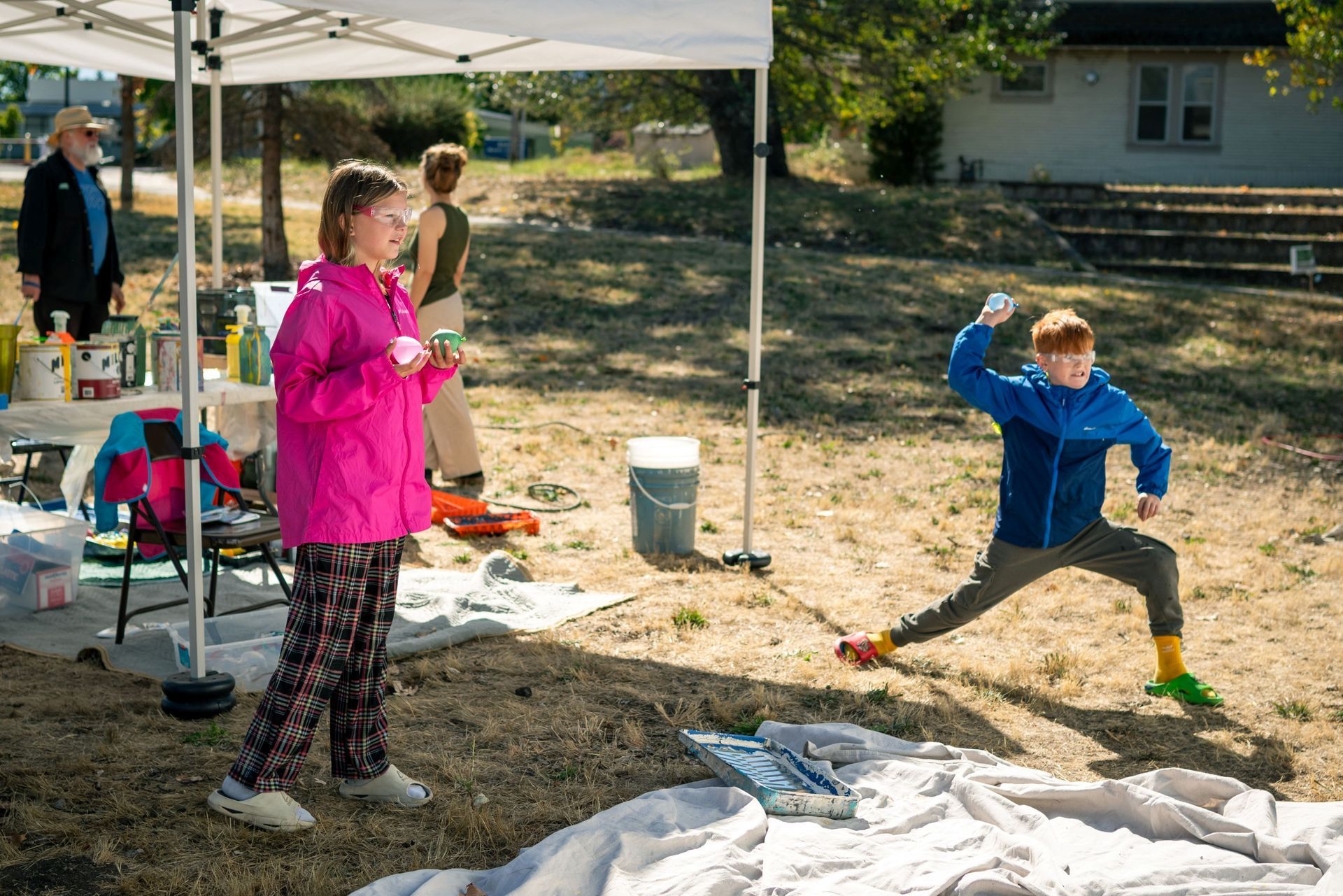 People outdoors under a canopy, one person in pink watching another in blue throwing a paint balloon
