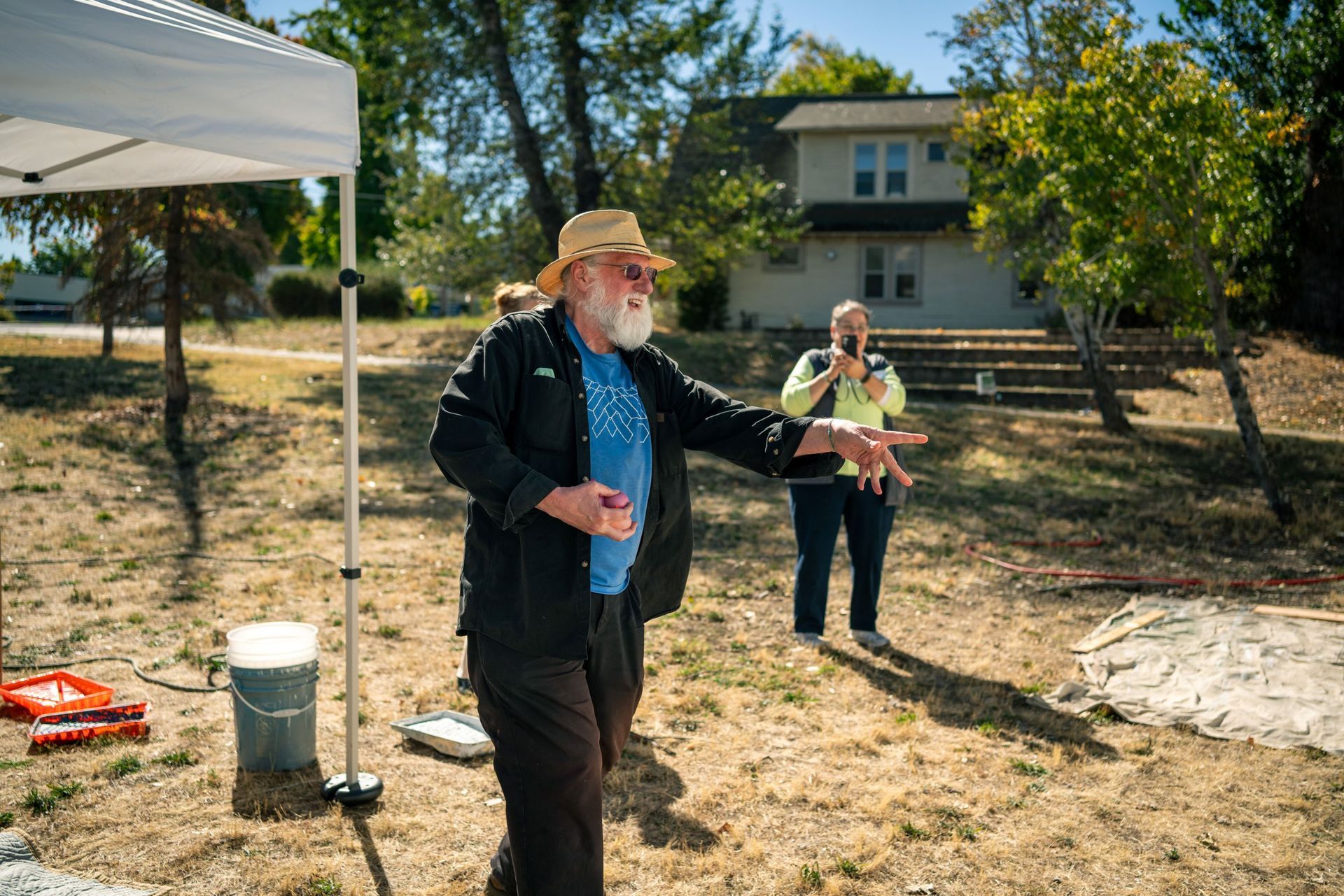 Man in a hat throwing a paint balloon outdoors near a woman holding a clipboard, under a canopy by a house and trees.