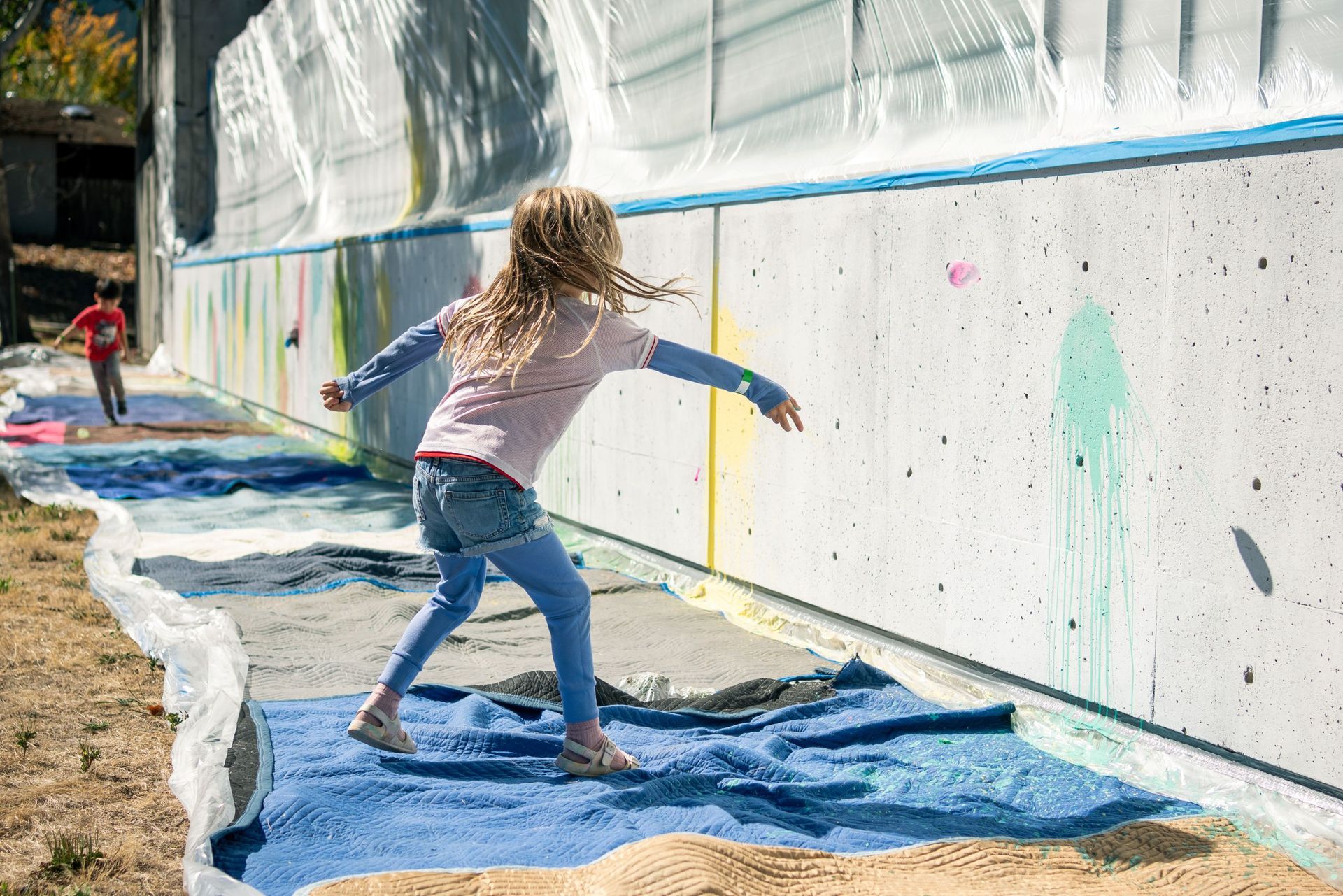 Child throwing paint balloon at a long wall outdoors with a blue tarp on the ground