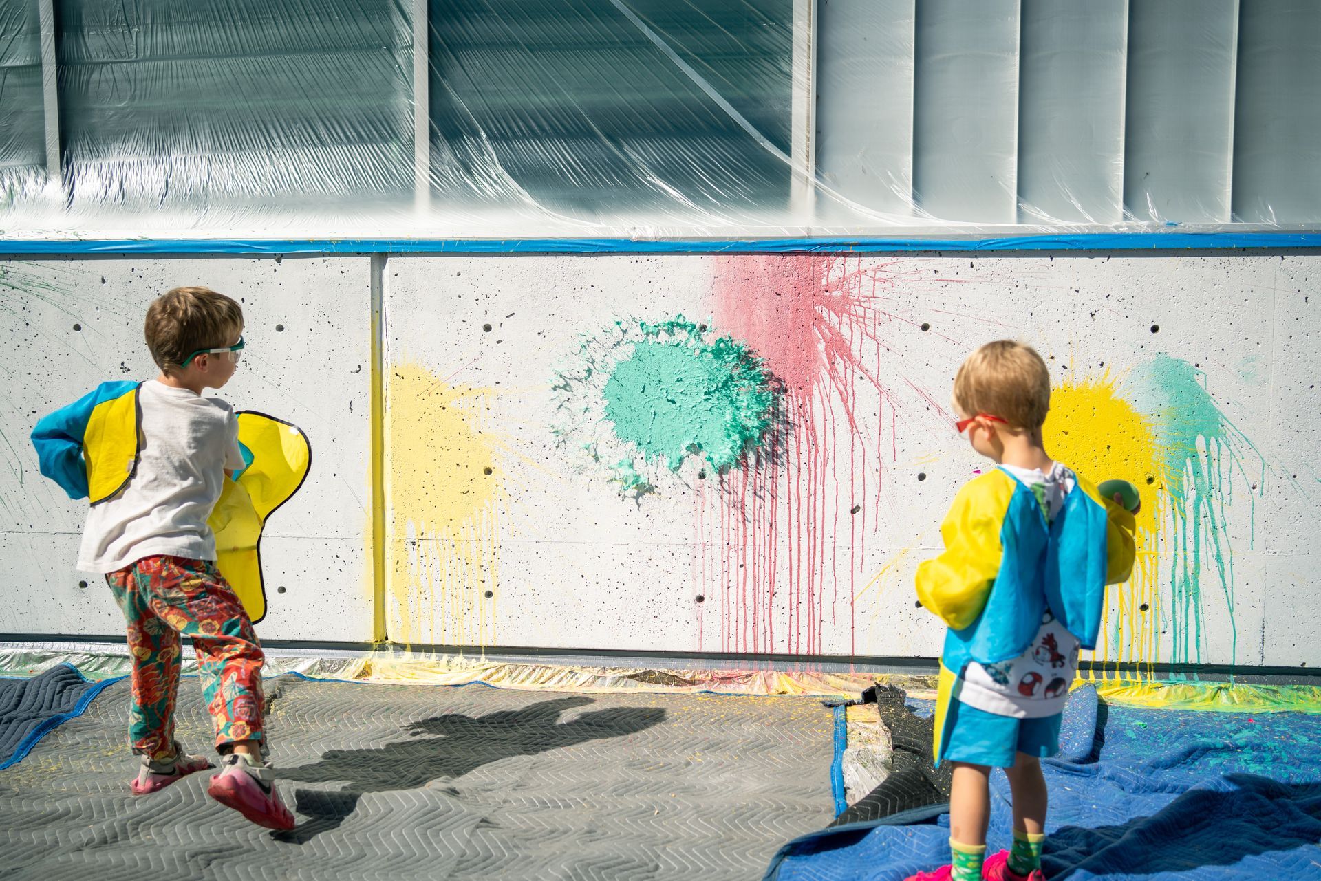 Two children throw paint balloons at a white wall creating bright splashes, outdoors.