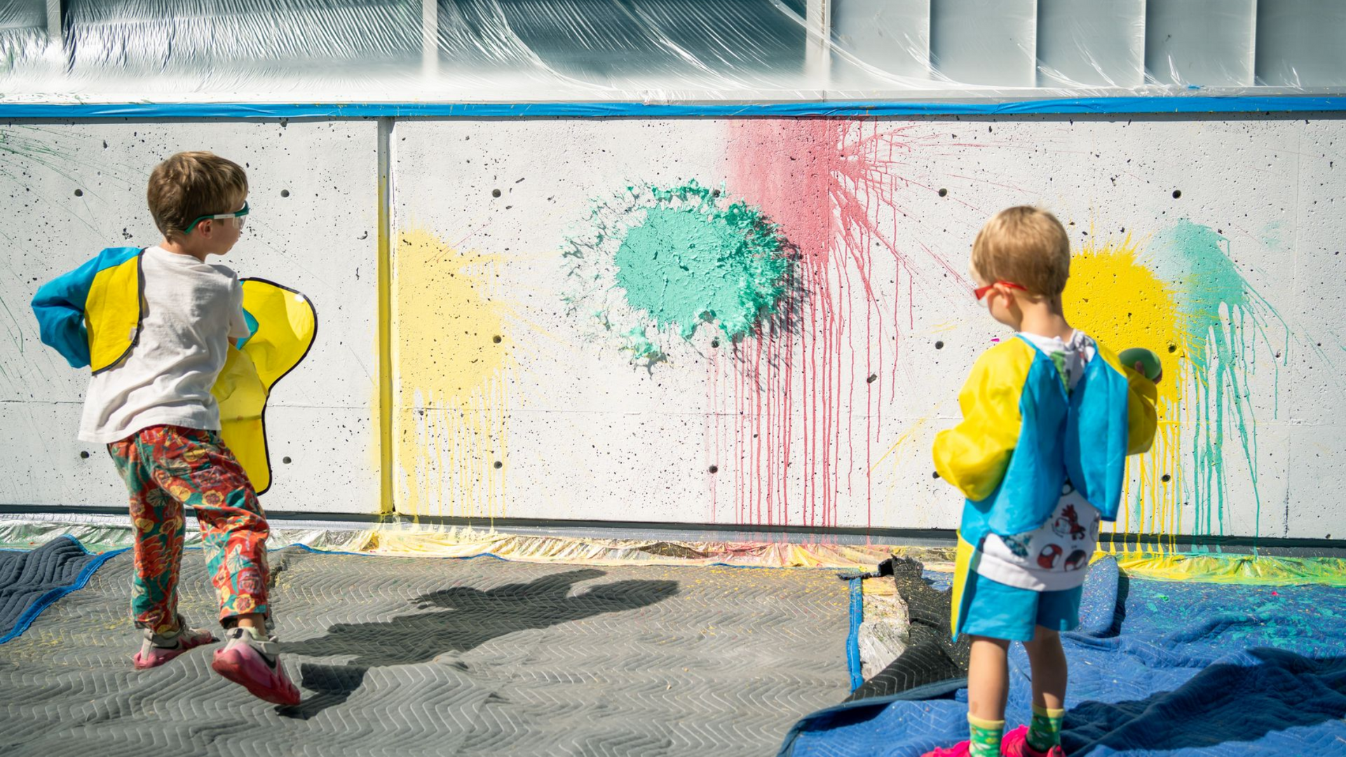 Two children create colorful splashes on a white outdoor wall with paint balloons