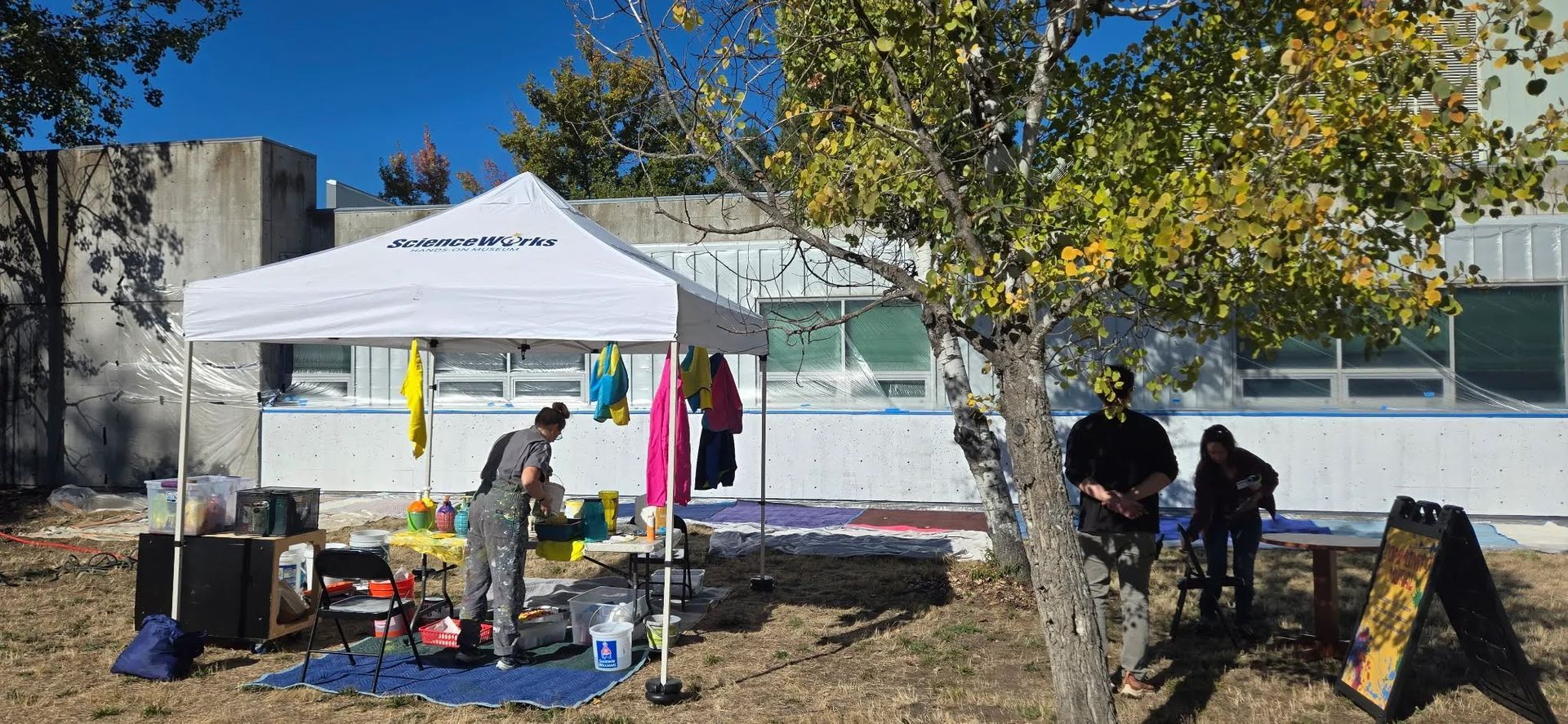 Outdoor white canopy with clothing, chairs, and people standing near a tree