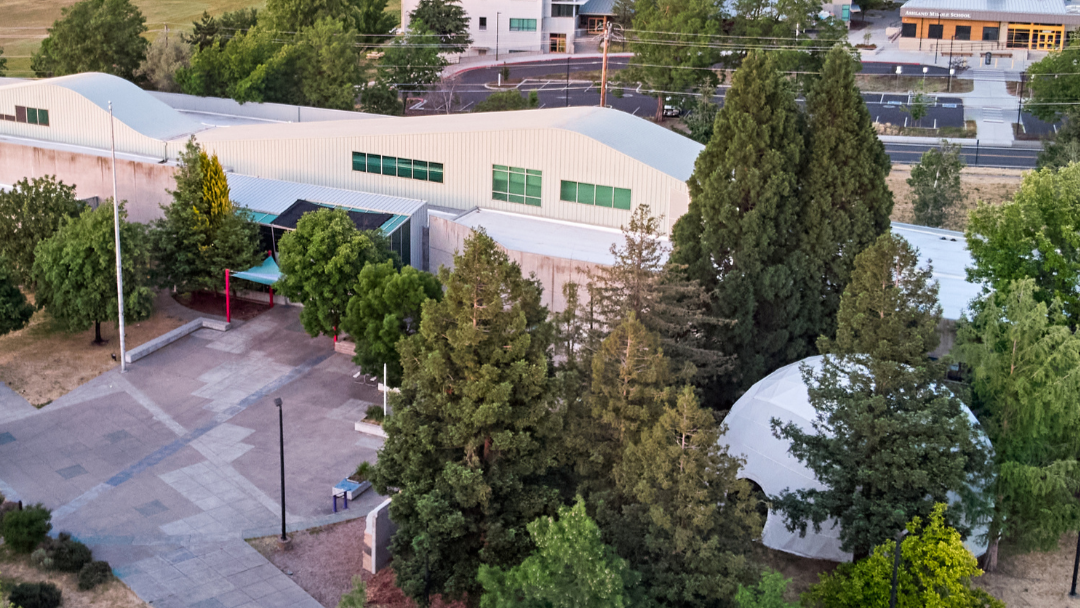Exterior view of building with a large dome structure and surrounding trees. Paved area in the foreground.