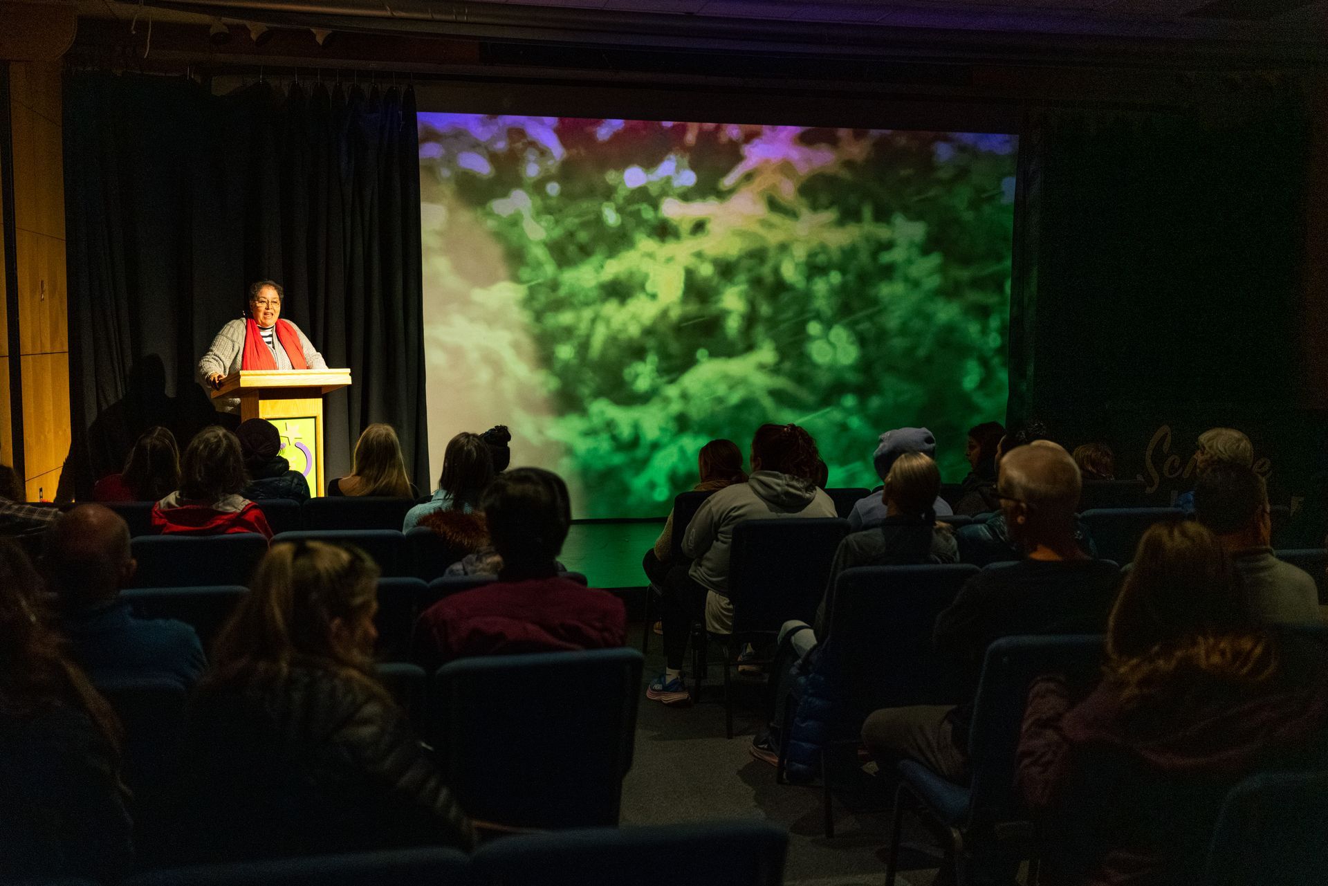 Speaker at podium addresses seated audience in a dim auditorium with a large nature image projected behind.