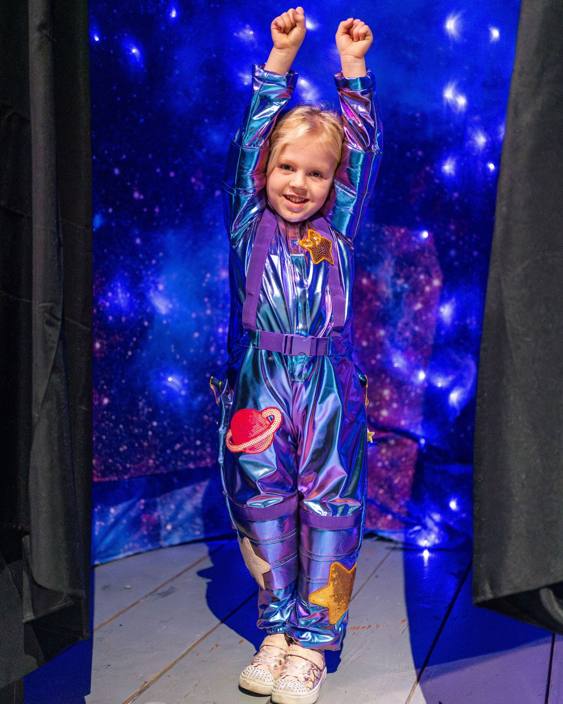 Child in a shiny blue astronaut costume with arms raised in front of a space-themed backdrop.