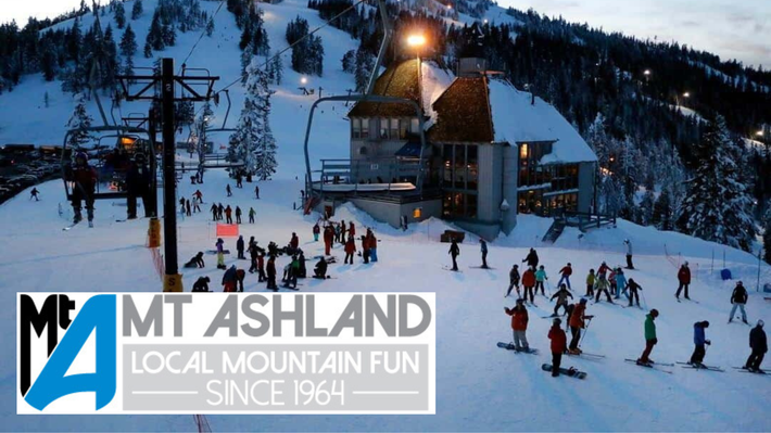 Skiers on a snowy mountain slope at Mt. Ashland, with a lodge and sign at dusk.