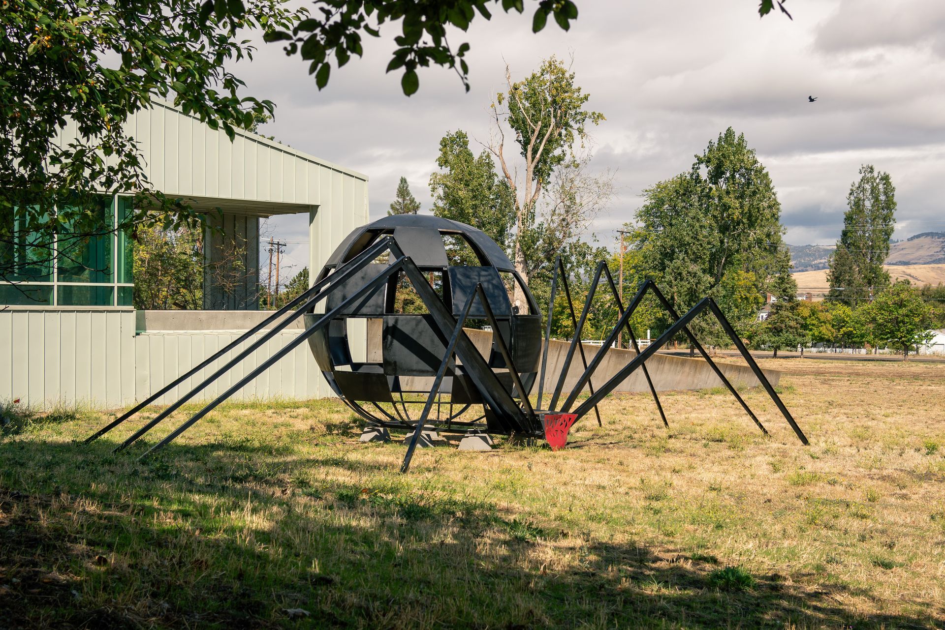 Black spider-like sculpture in a grassy field beside a building, with trees in the background