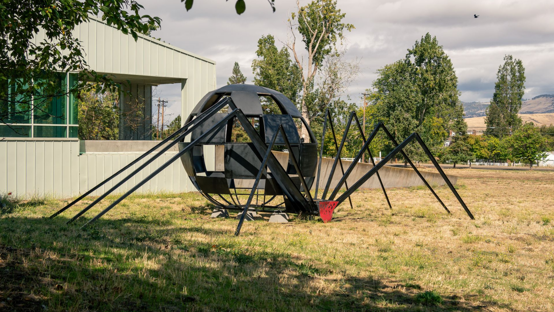 Black geometric spider sculpture on grass near a building, with trees and mountains in the background