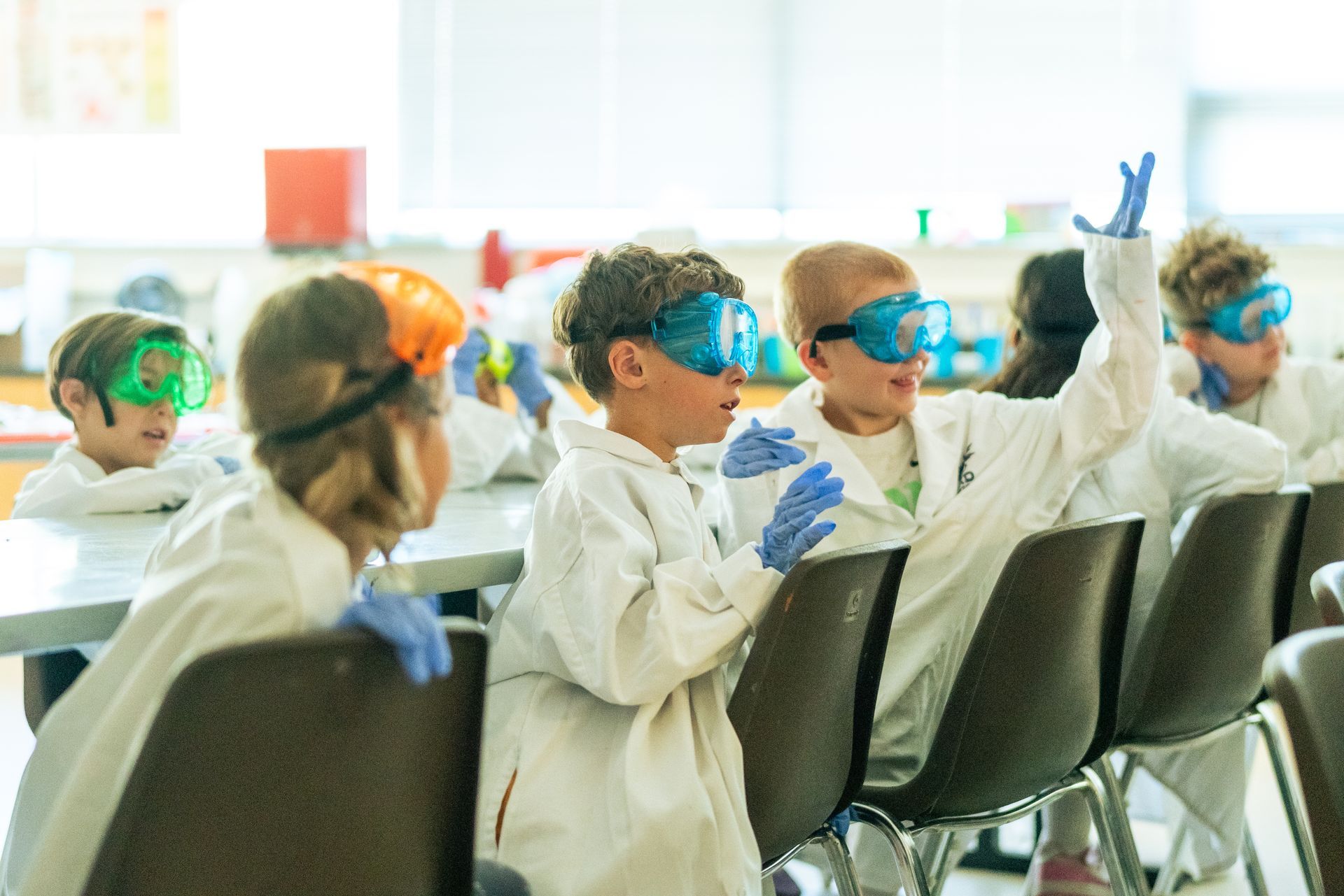 Children in lab coats, safety goggles, and gloves; one child raises their hand in a science classroom.