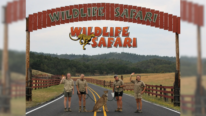 People posing at the Wild Life Safari entrance arch on a road with fences and forested hills.