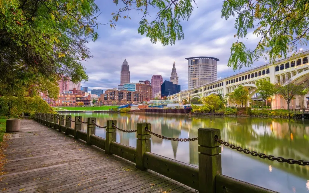 Cleveland, Ohio skyline along a river with bridge, greenery, and cloudy sky.