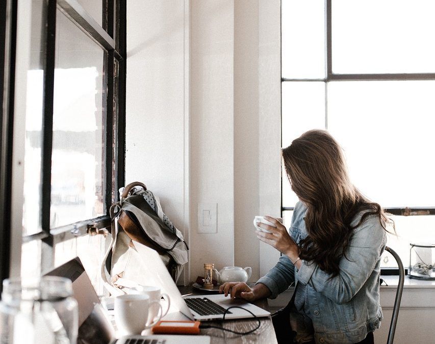 Woman seated at desk near window, holding mug, using laptop.