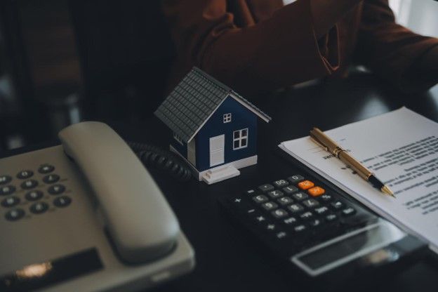 Miniature house model, calculator, paperwork, pen, and phone on a desk, representing real estate or home finances.