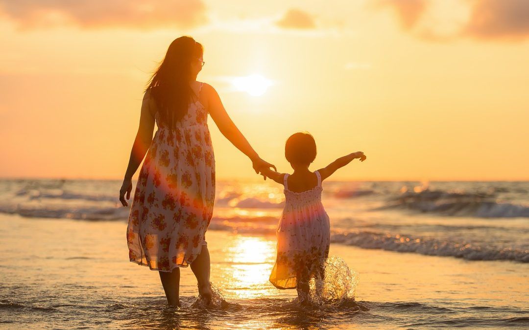 Woman and child holding hands, walking into ocean at sunset.