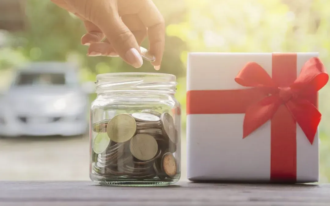 Hand dropping coin into jar of coins next to a wrapped gift and a car.