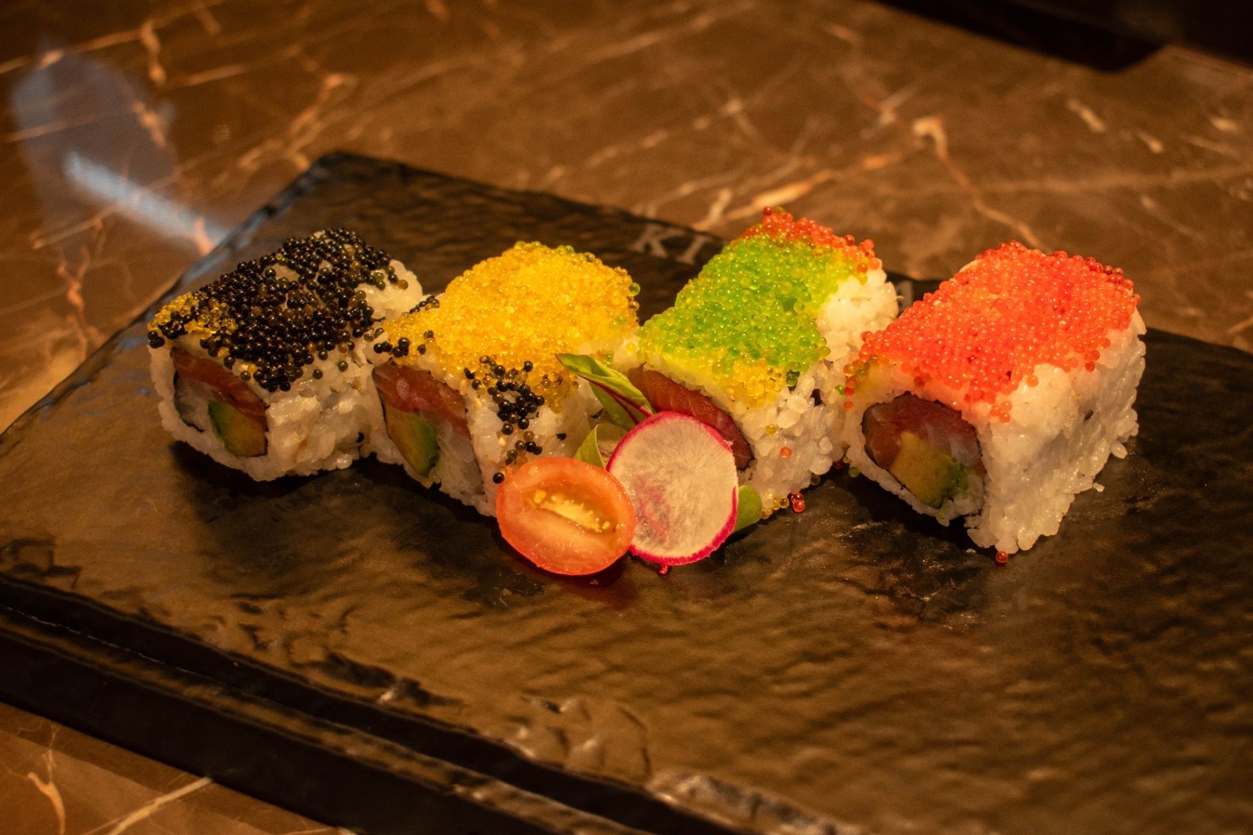 Colorful sushi rolls topped with black, yellow, green, and red roe on a black plate