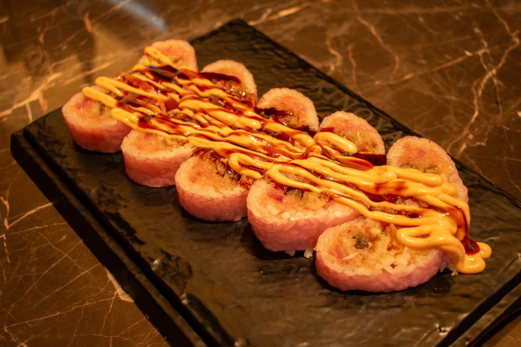 Sushi rolls topped with orange sauce on a black serving board, arranged on a marble countertop