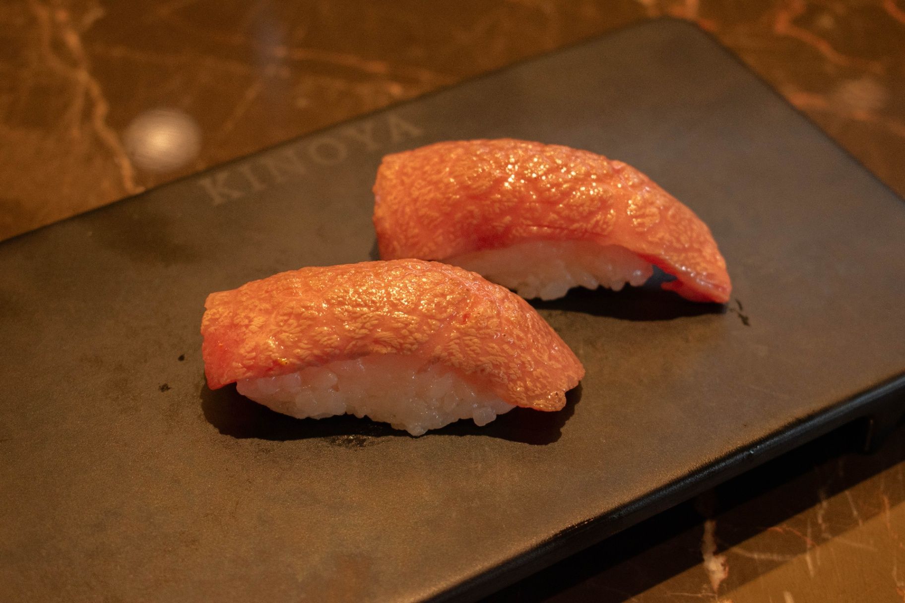 Two pieces of sushi on a black plate, with pink fish atop white rice, on a wooden table.