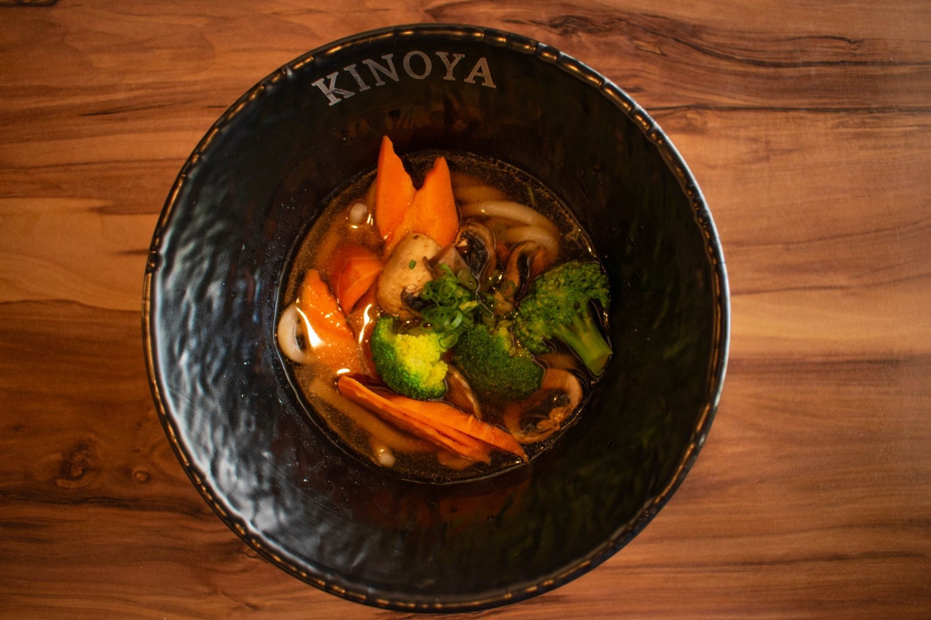 Black bowl of ramen with orange broth, broccoli, carrots, and mushrooms on a wooden table.
