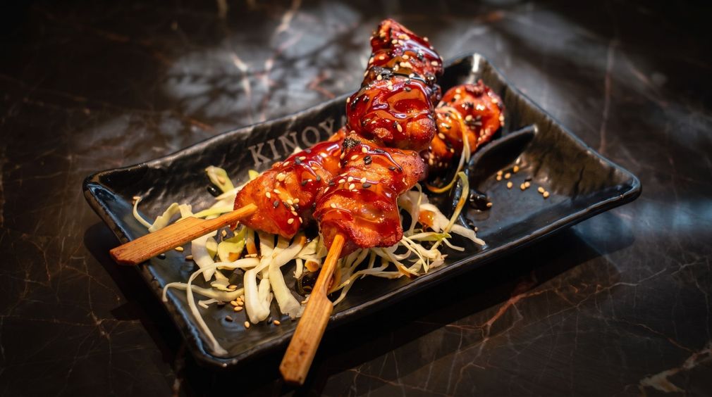 Skewered glazed chicken over noodles on a black plate, with chopsticks and garnish on a dark table