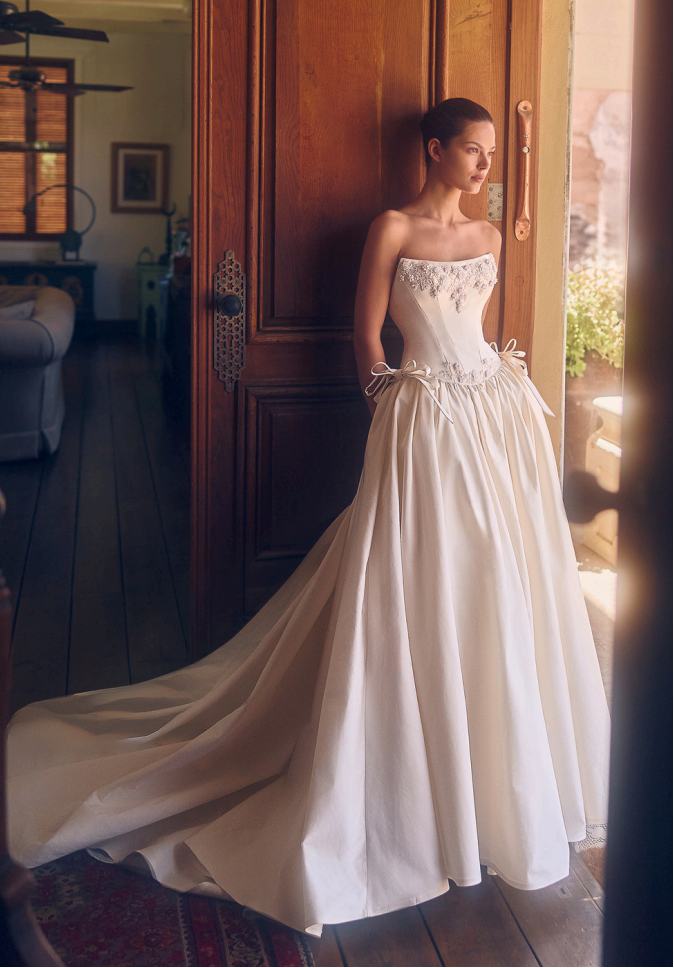 Woman in a white wedding dress stands by a gold cabinet, facing left.