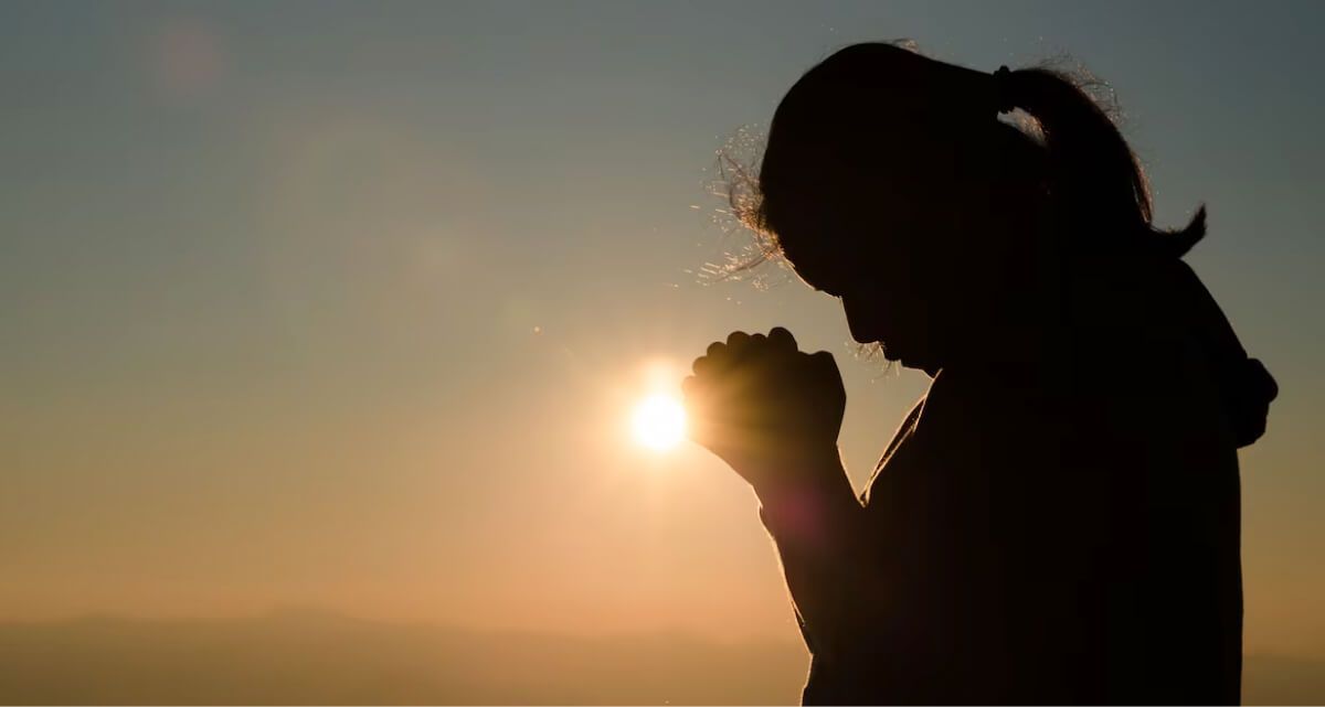 A woman is praying with her hands folded in front of the sun.