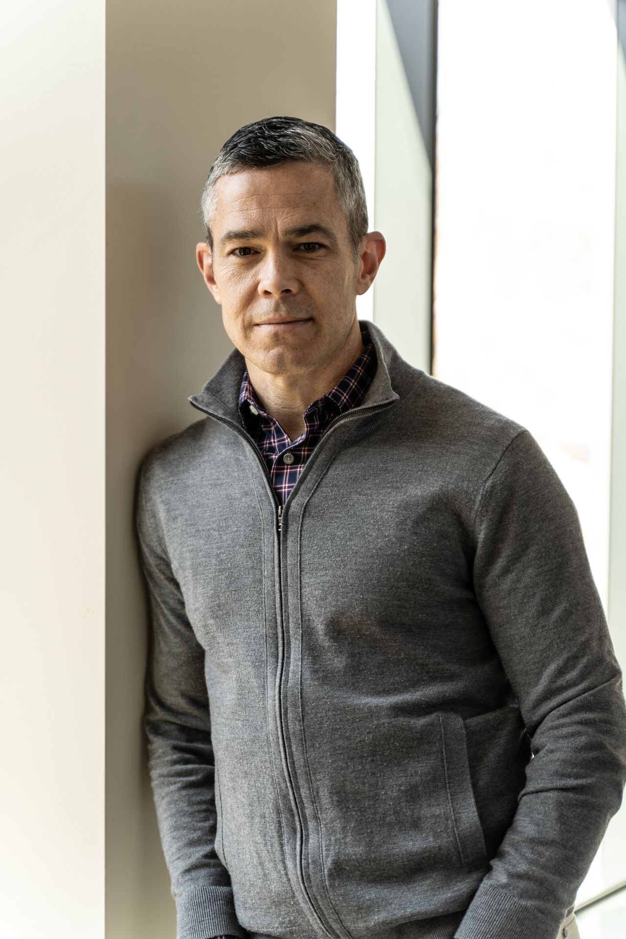 Man standing by a bright window, wearing a gray zip-up sweater and patterned shirt.