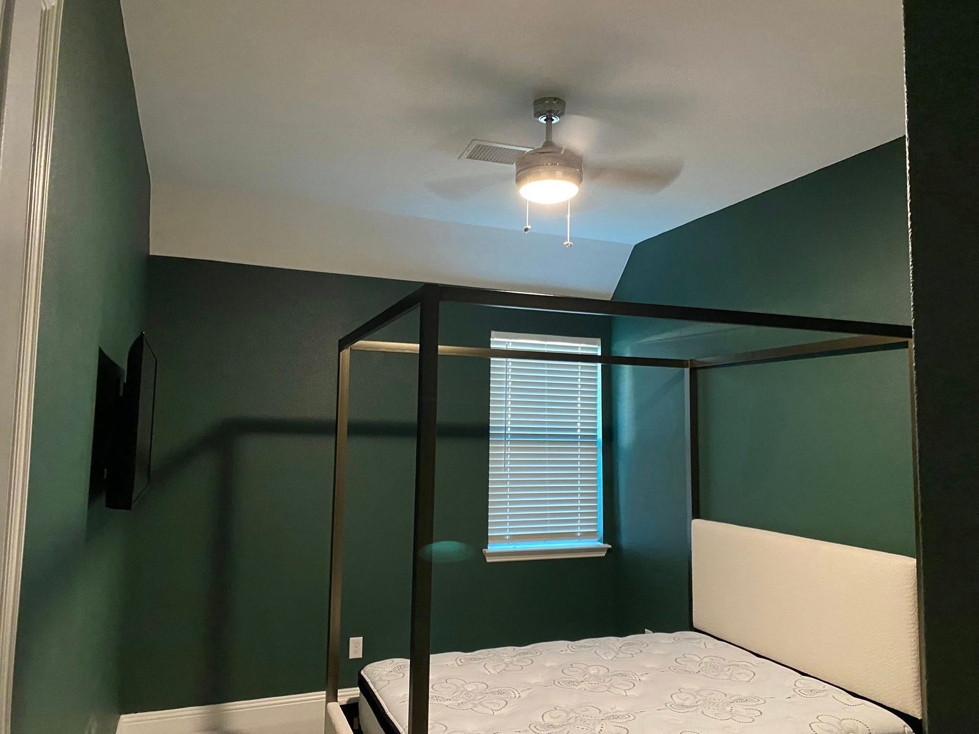 Green-walled bedroom with canopy bed, window, ceiling fan, and mounted TV.