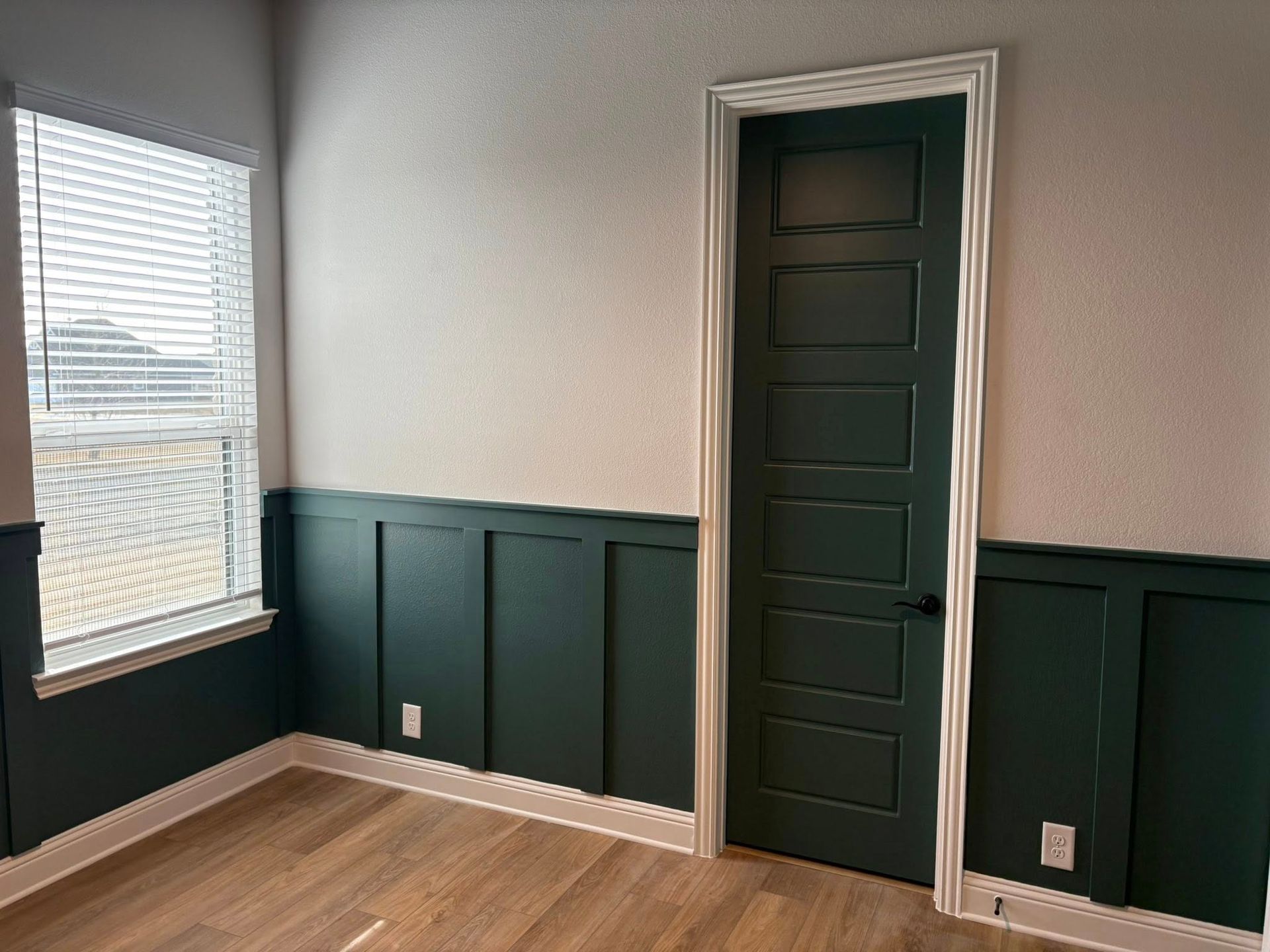 Room with dark green wainscoting and door, white walls, wood floor, window with blinds.