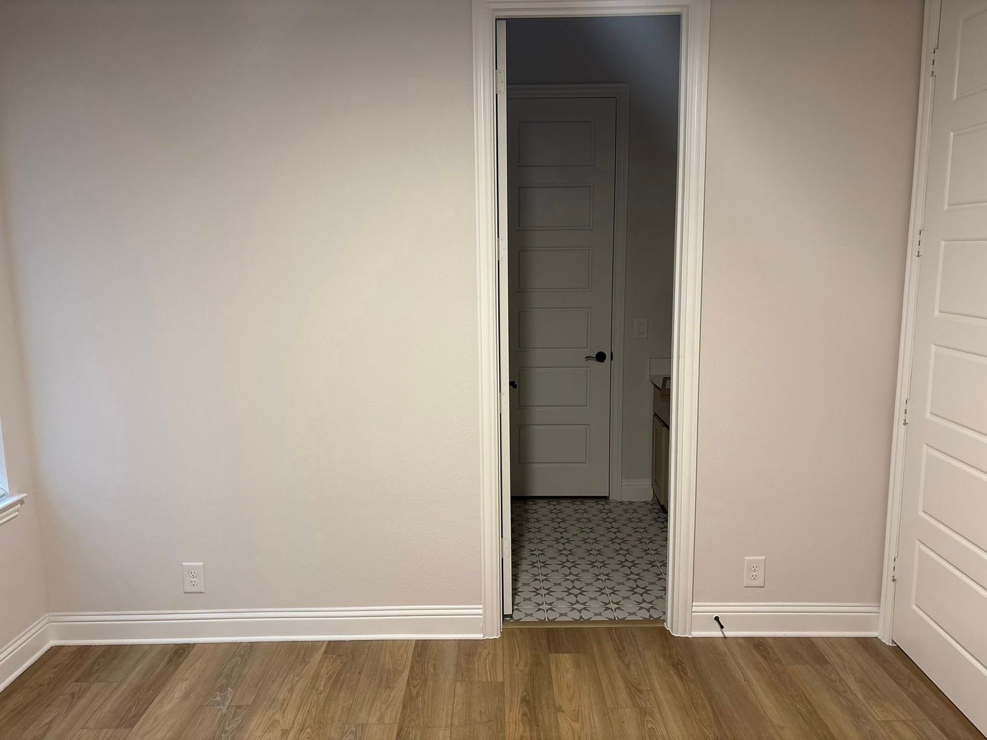 Empty room with light-colored walls and wooden floors. An open doorway leads to a smaller room with a patterned floor.