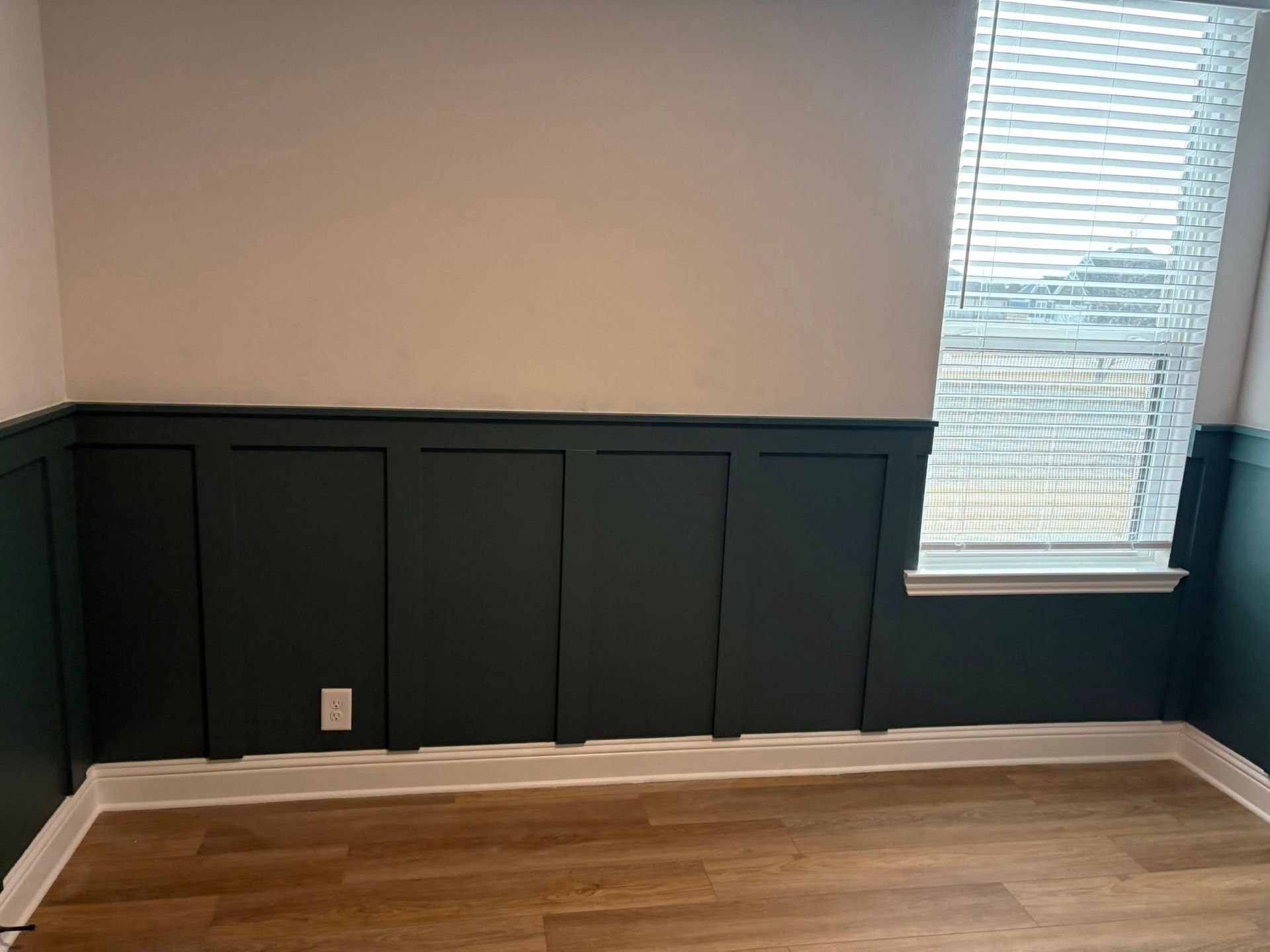 Room with dark green wainscoting and tan upper wall, next to a window with blinds.