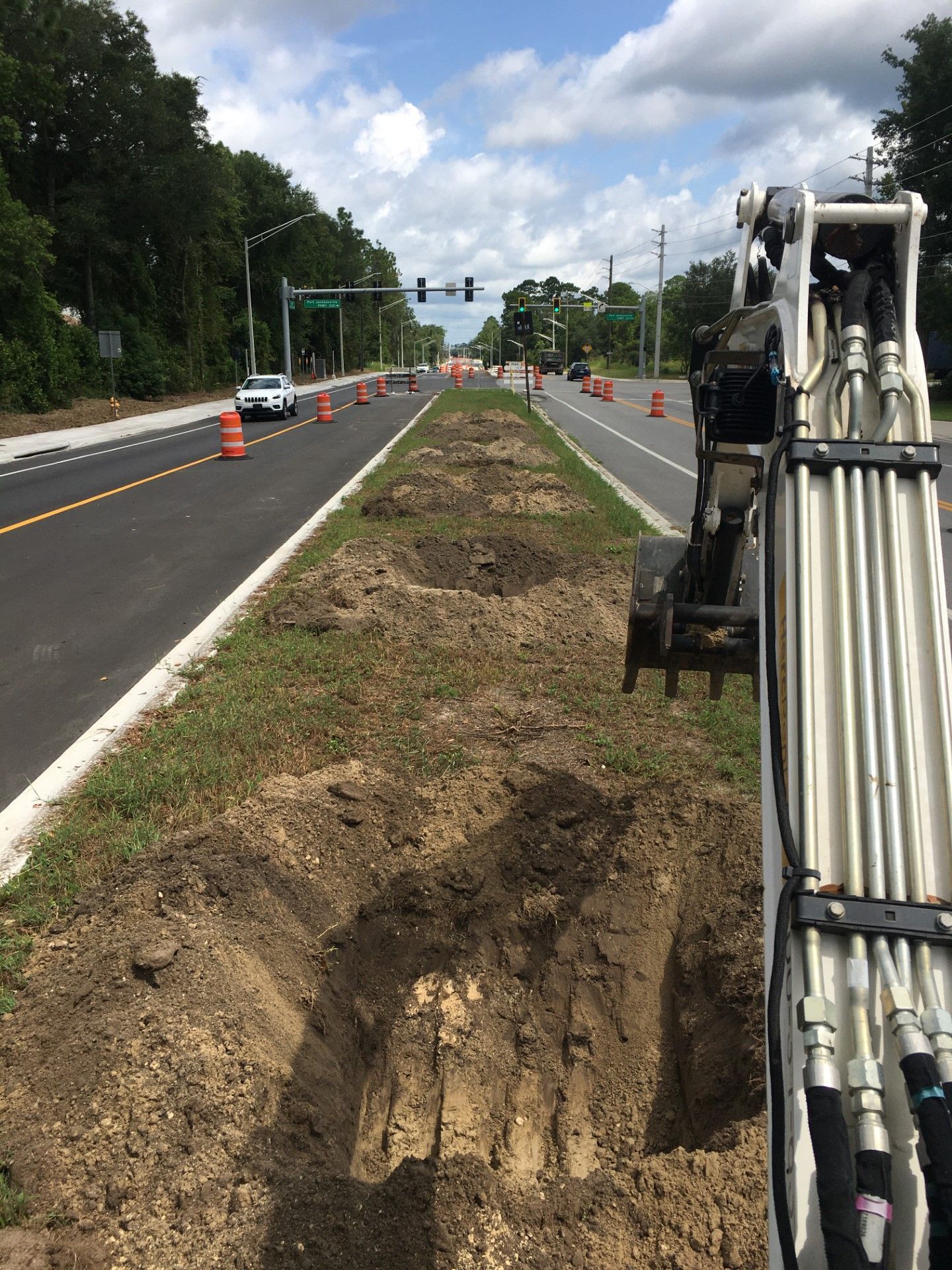 Road construction: Excavations in median, orange cones, excavator arm in view, clear sky.