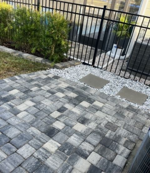 Paver patio with varying gray stones, next to a black fence and green shrubbery.