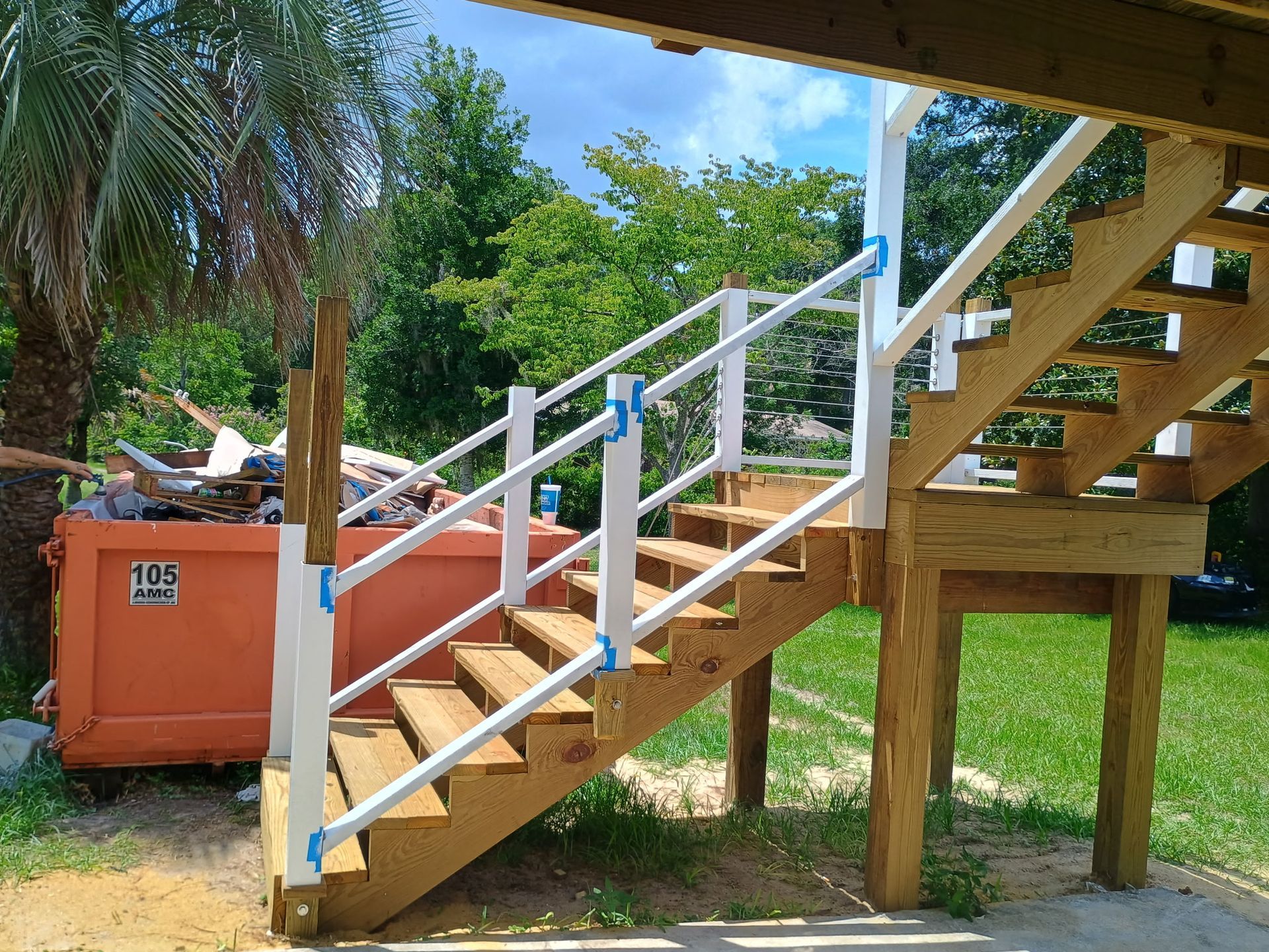 Wooden staircase with white railings, leading up to a deck, with a dumpster in the background.