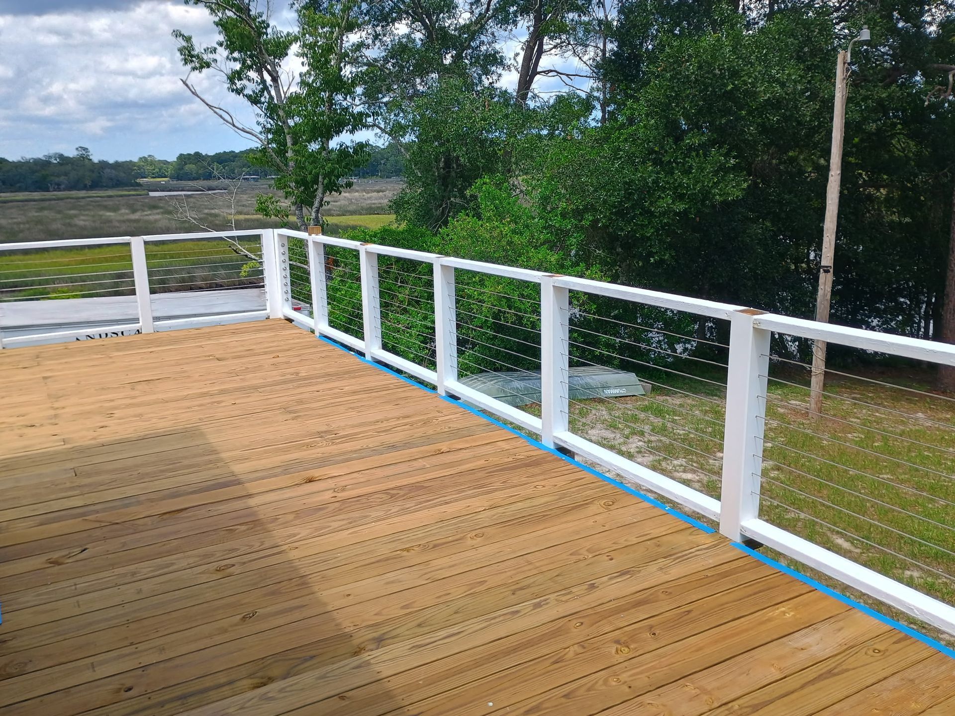 Wooden deck with white railing and cable, overlooking a marshy landscape with trees.
