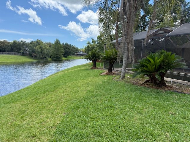 Lush green lawn slopes down to a calm lake. Trees line the bank, with a screened-in porch visible to the right.